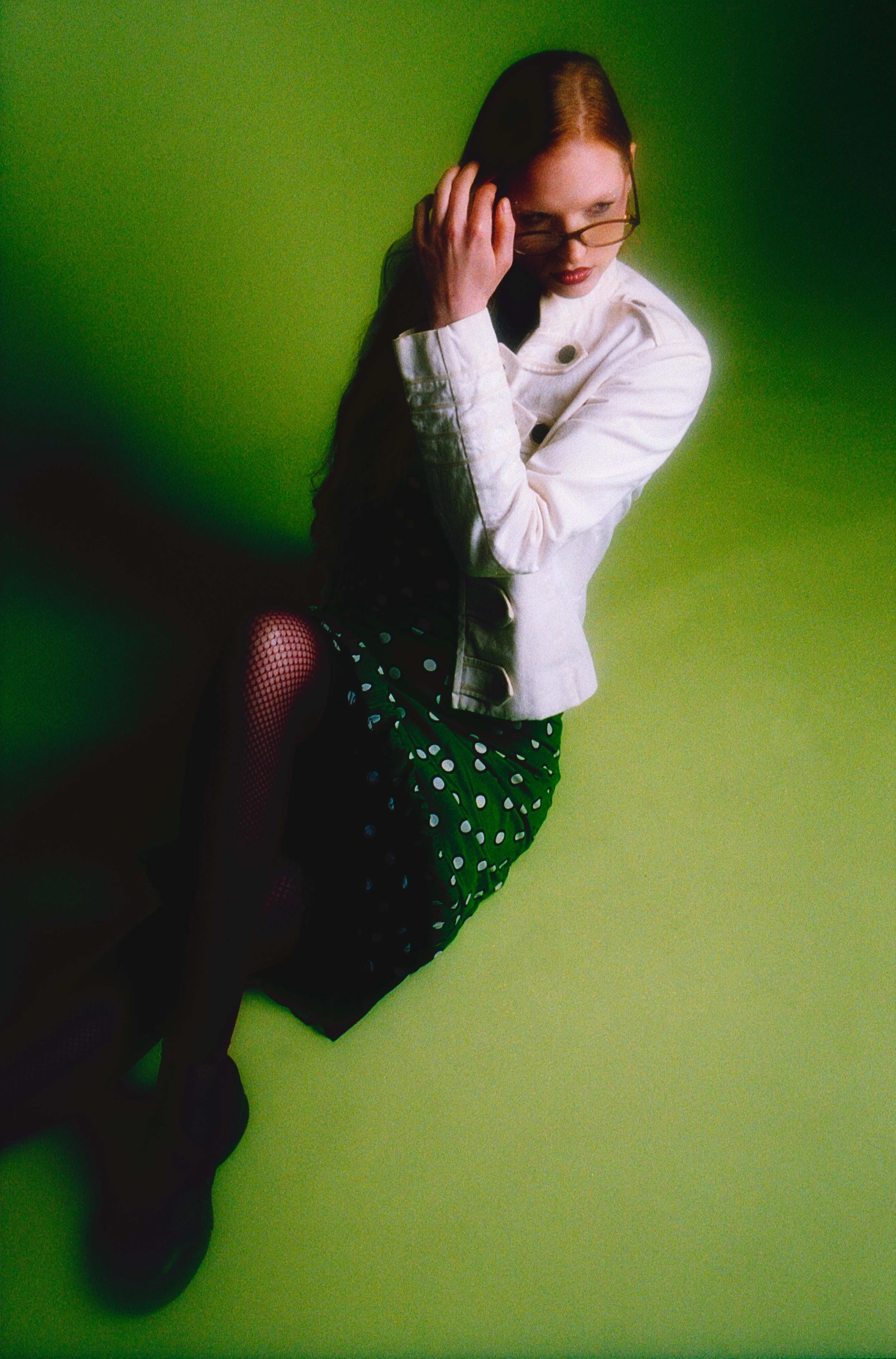 Model with glasses in a white jacket and polka dot skirt sitting on a deep green floor. Vintáááge May by Hartenjager.