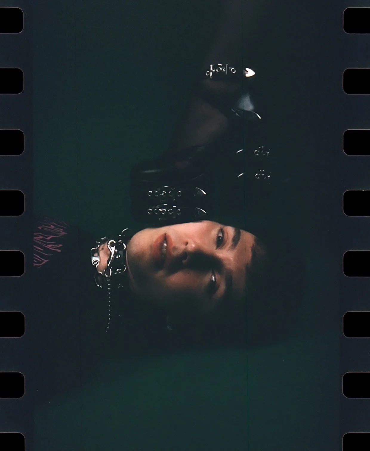 Dark film frame of a face lying sideways with a studded collar and chains in shadow. Dominance by Hartenjager.