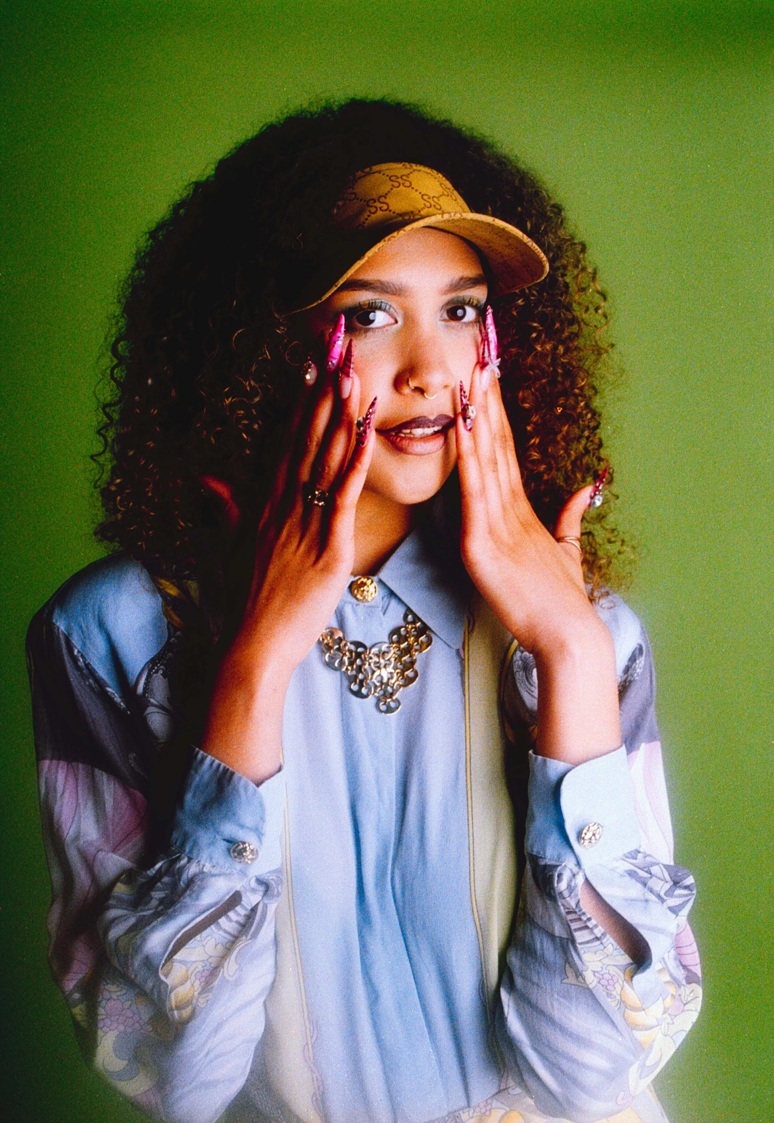 Curly-haired model in a cap and pale blue shirt, hands on her cheeks, mouth open. Vintáááge May by Hartenjager.