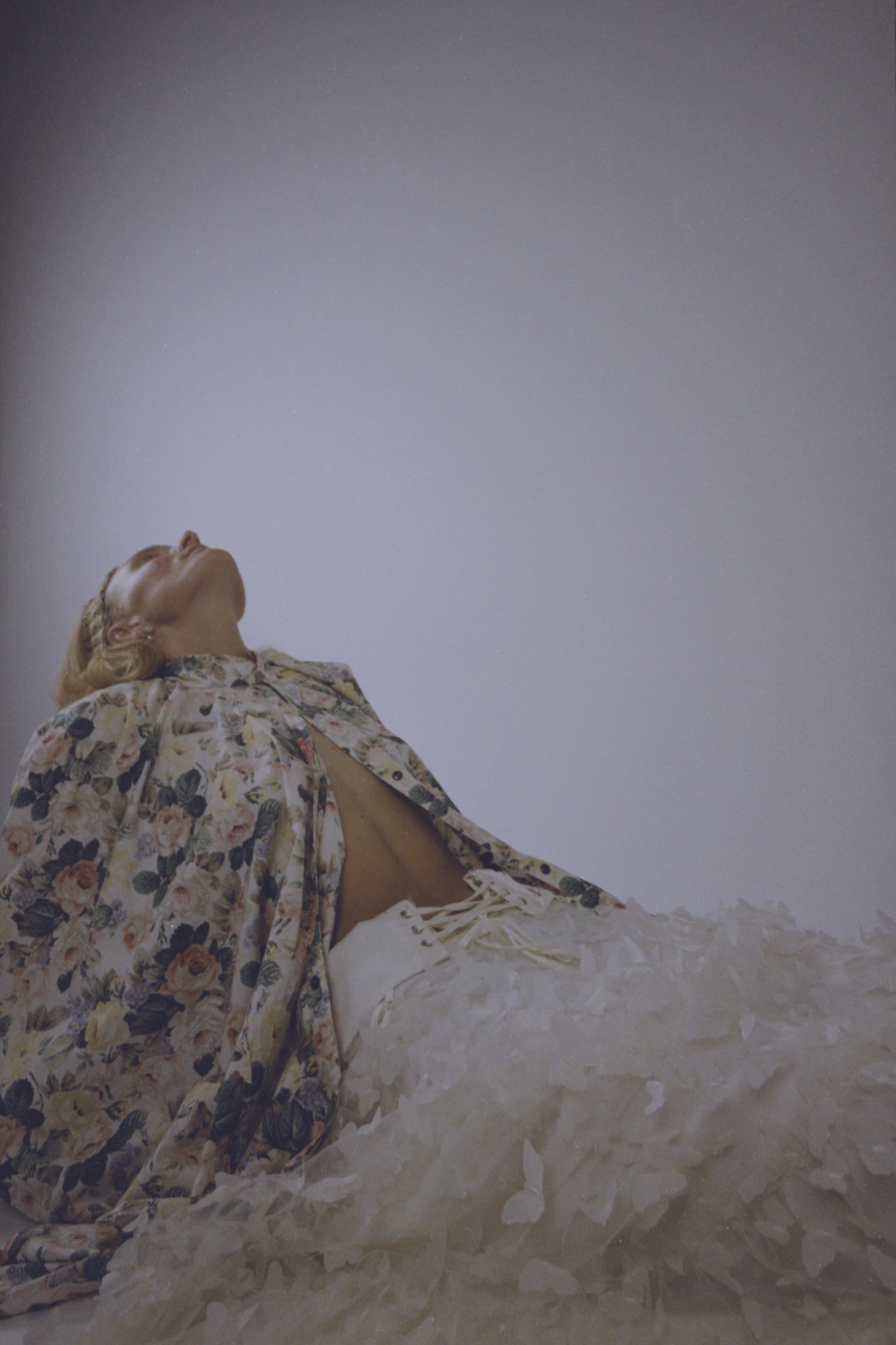 Model lying back in a floral coat and white tulle skirt, face turned up toward the light. Vintáááge January by Hartenjager.