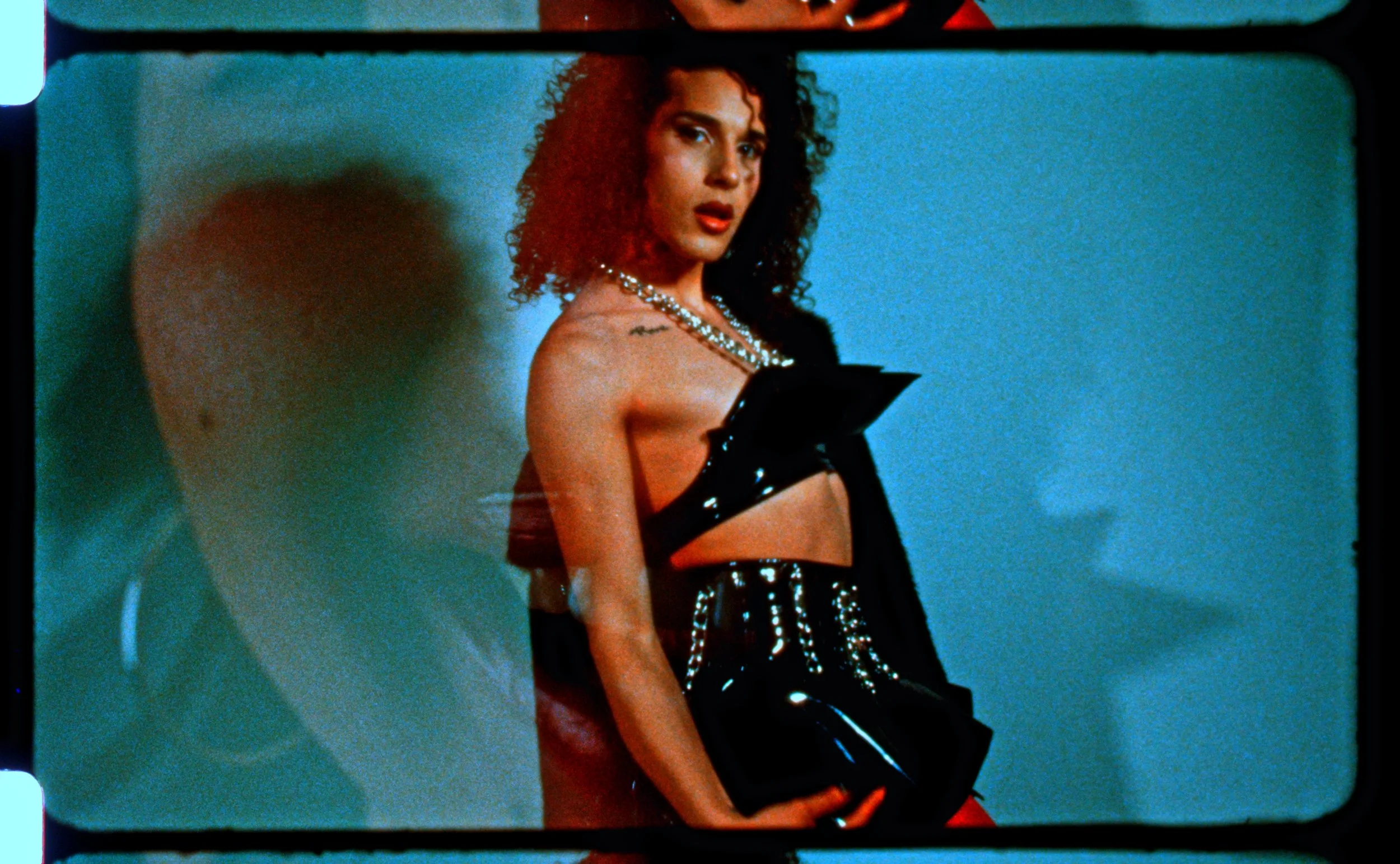 16mm frame of a curly-haired figure in a black harness against a blue backdrop. Still from Intrusive Thots, 16mm film by Hartenjager.