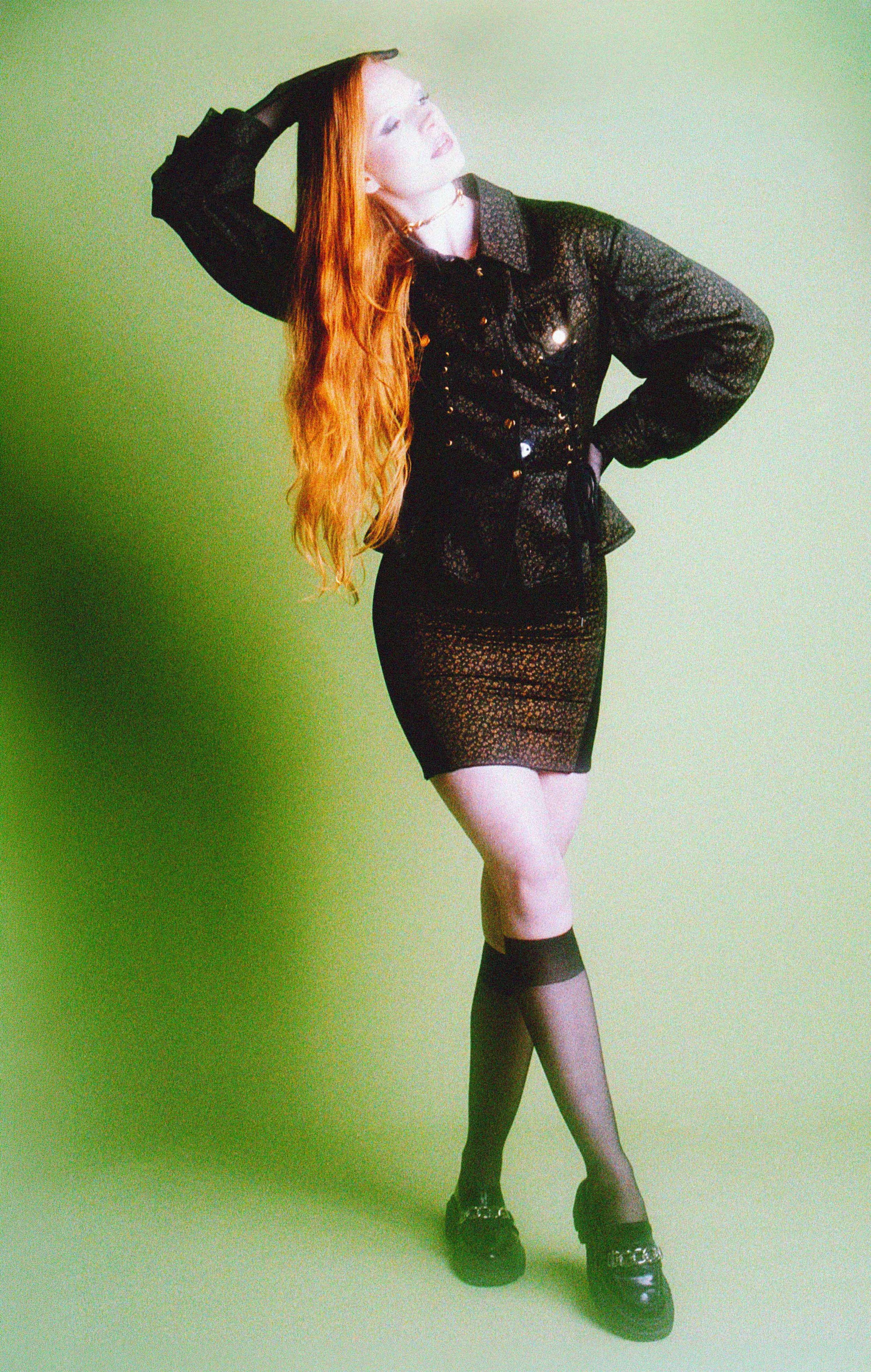 Red-haired model in a black jacket and mini skirt with knee socks, posing against a green backdrop. Vintáááge May by Hartenjager.