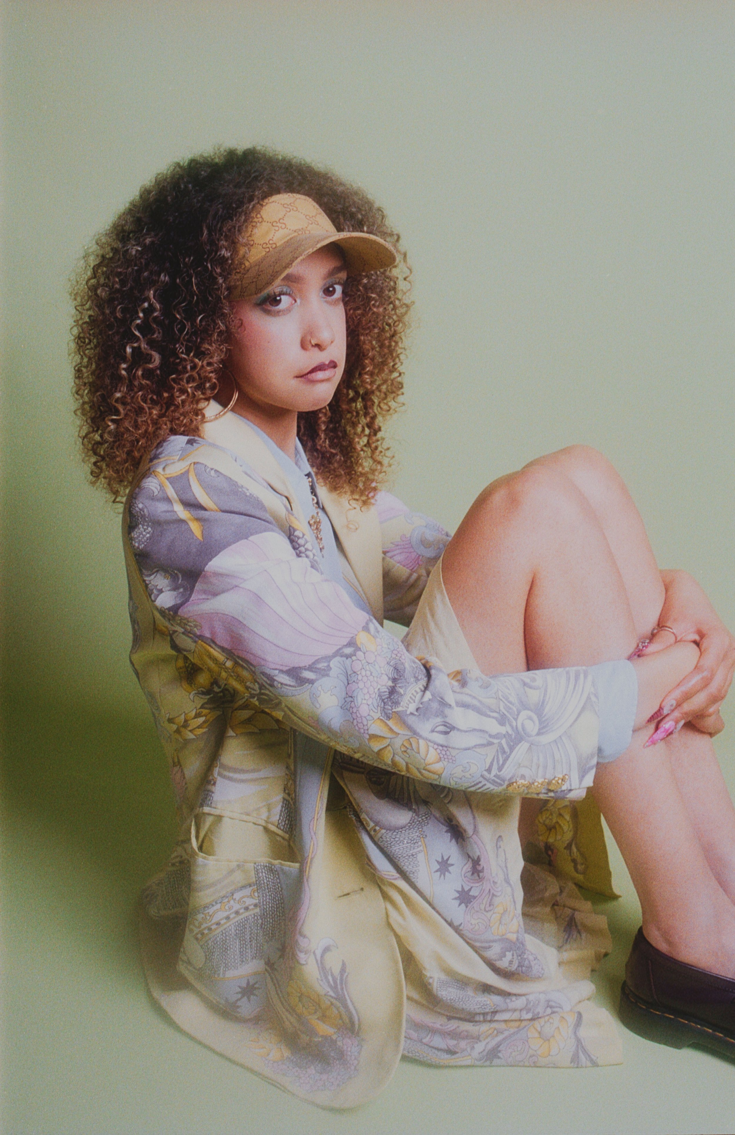 Curly-haired model in a cap and printed jacket sitting with knees up against a pale green wall. Vintáááge May by Hartenjager.