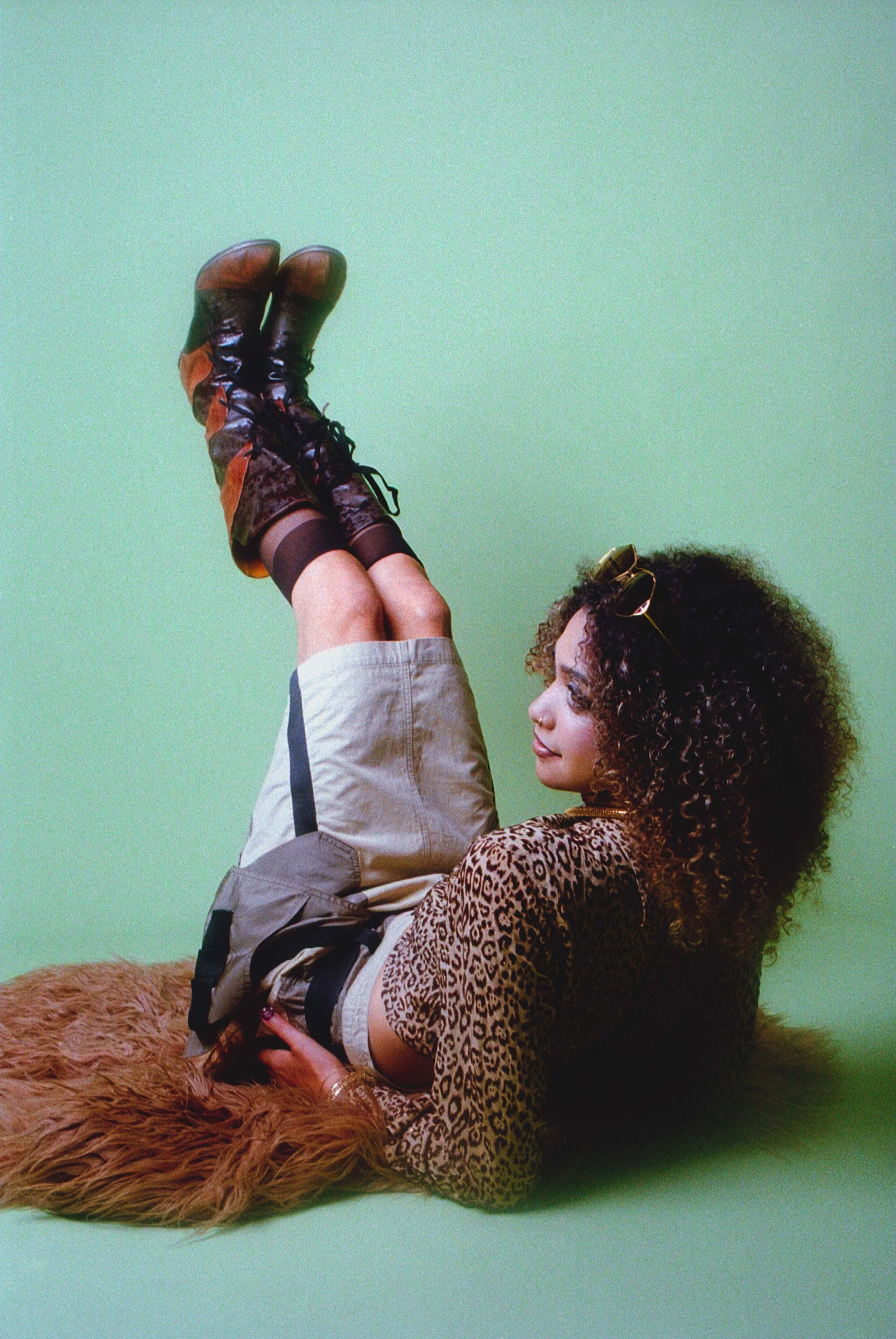 Curly-haired model lying on a fur rug with boots up in the air, in a leopard print jacket. Vintáááge May by Hartenjager.
