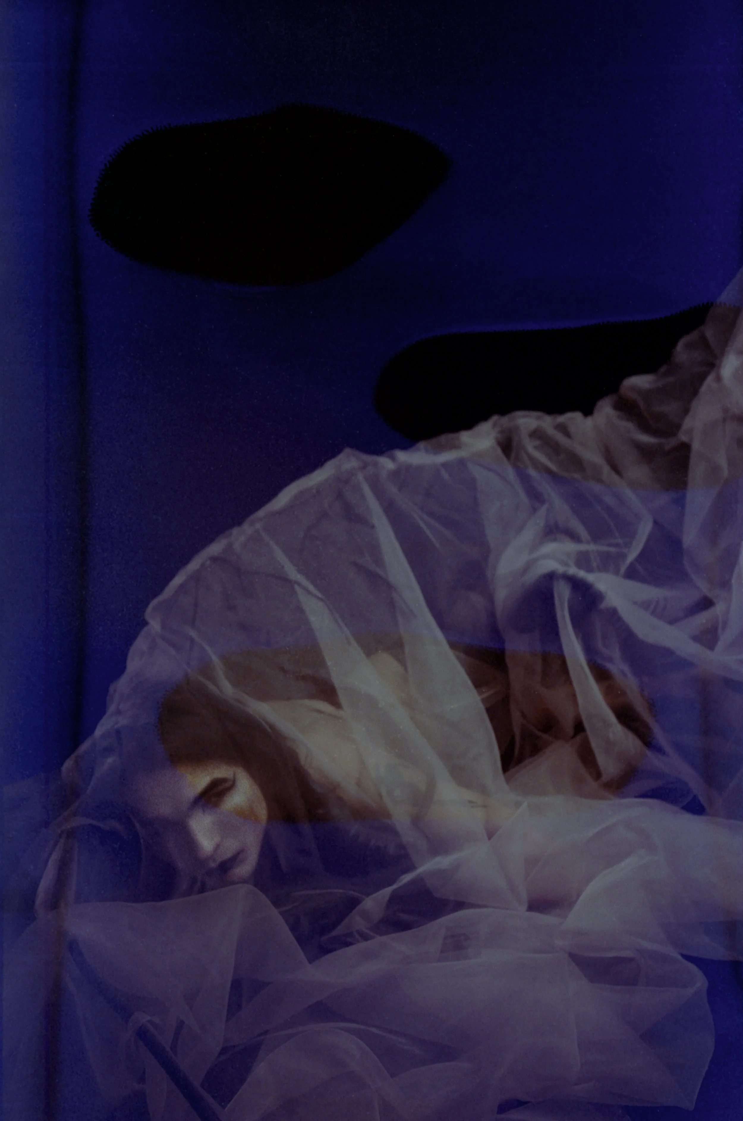 Figure lying under translucent fabric in deep blue light, partly obscured by dark film damage. By Hartenjager.