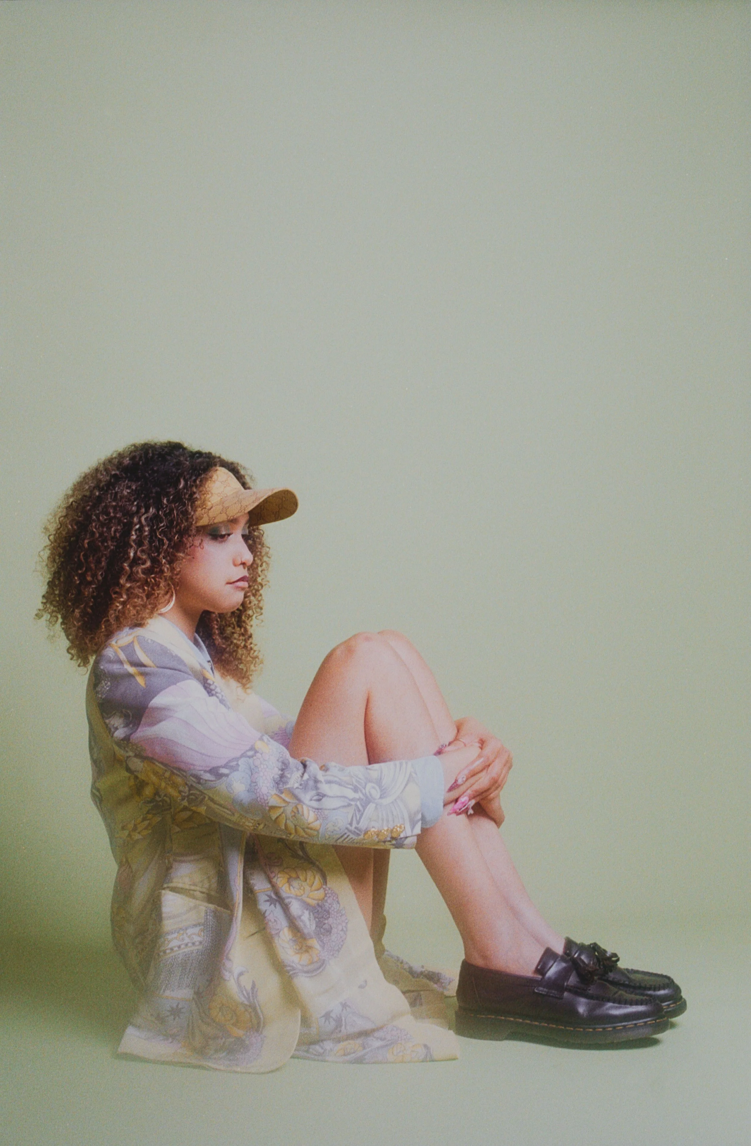 Curly-haired model in a cap sitting on the floor in a printed jacket and black shoes. Vintáááge May by Hartenjager.
