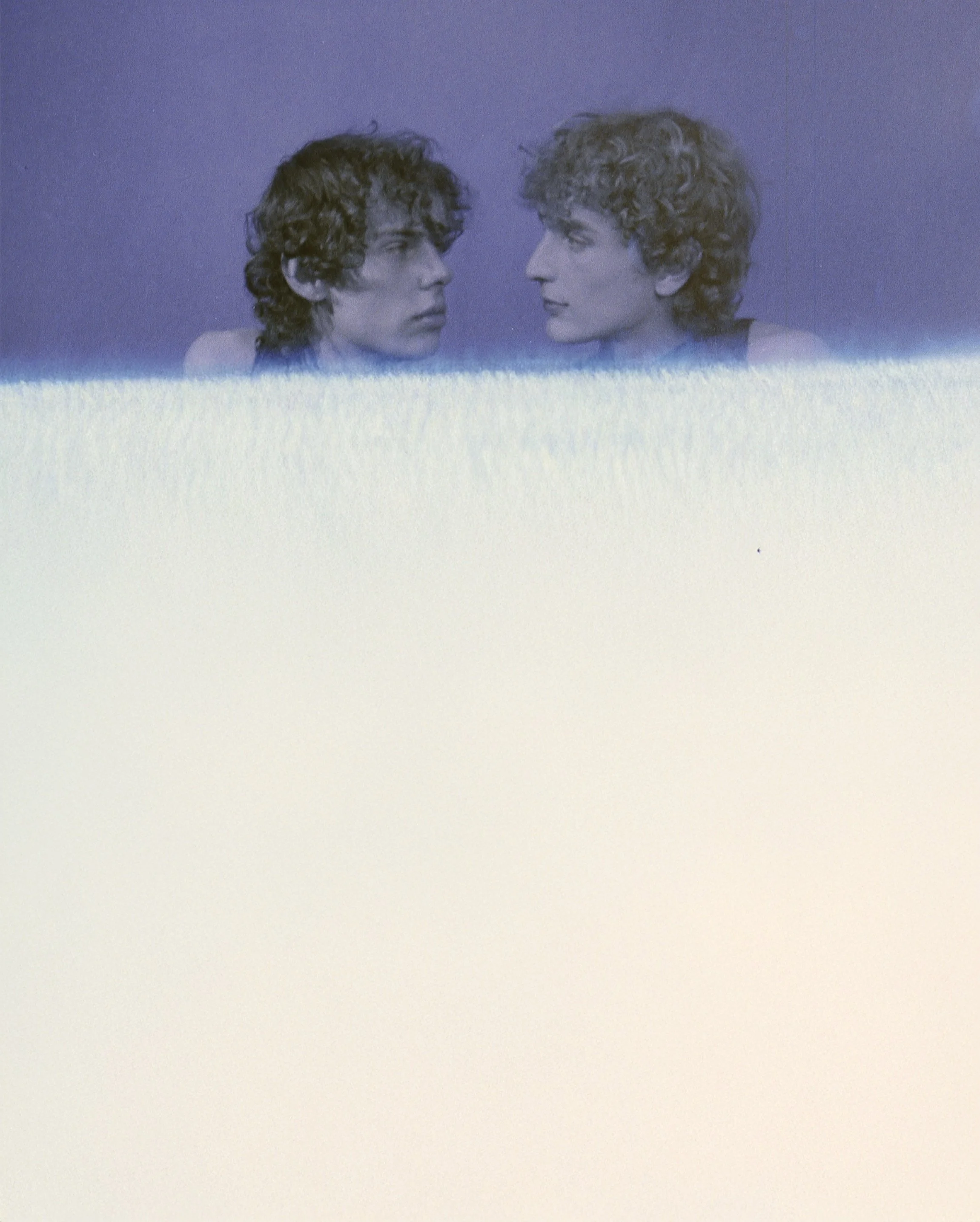 Two young men with curly hair facing each other above a washed-out light leak. Geminus by Hartenjager.