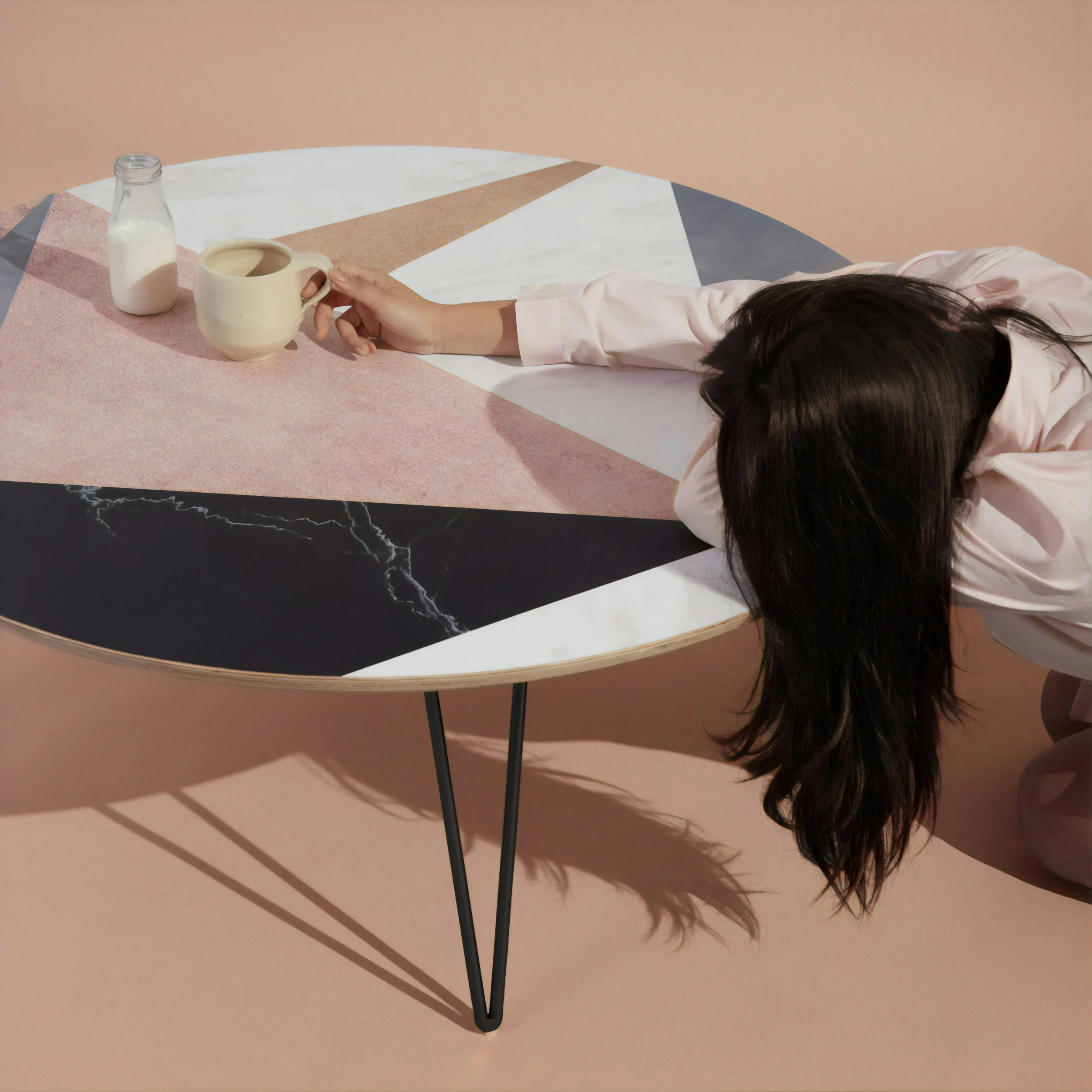 A photo of a woman with long, dark hair covering her face, laying over the top of a table. One hand barely holds the edge of a coffee a cup, a bottle of creamer beside it. The image evokes a sense of exhaustion and burnout. Therapy for High Achievers