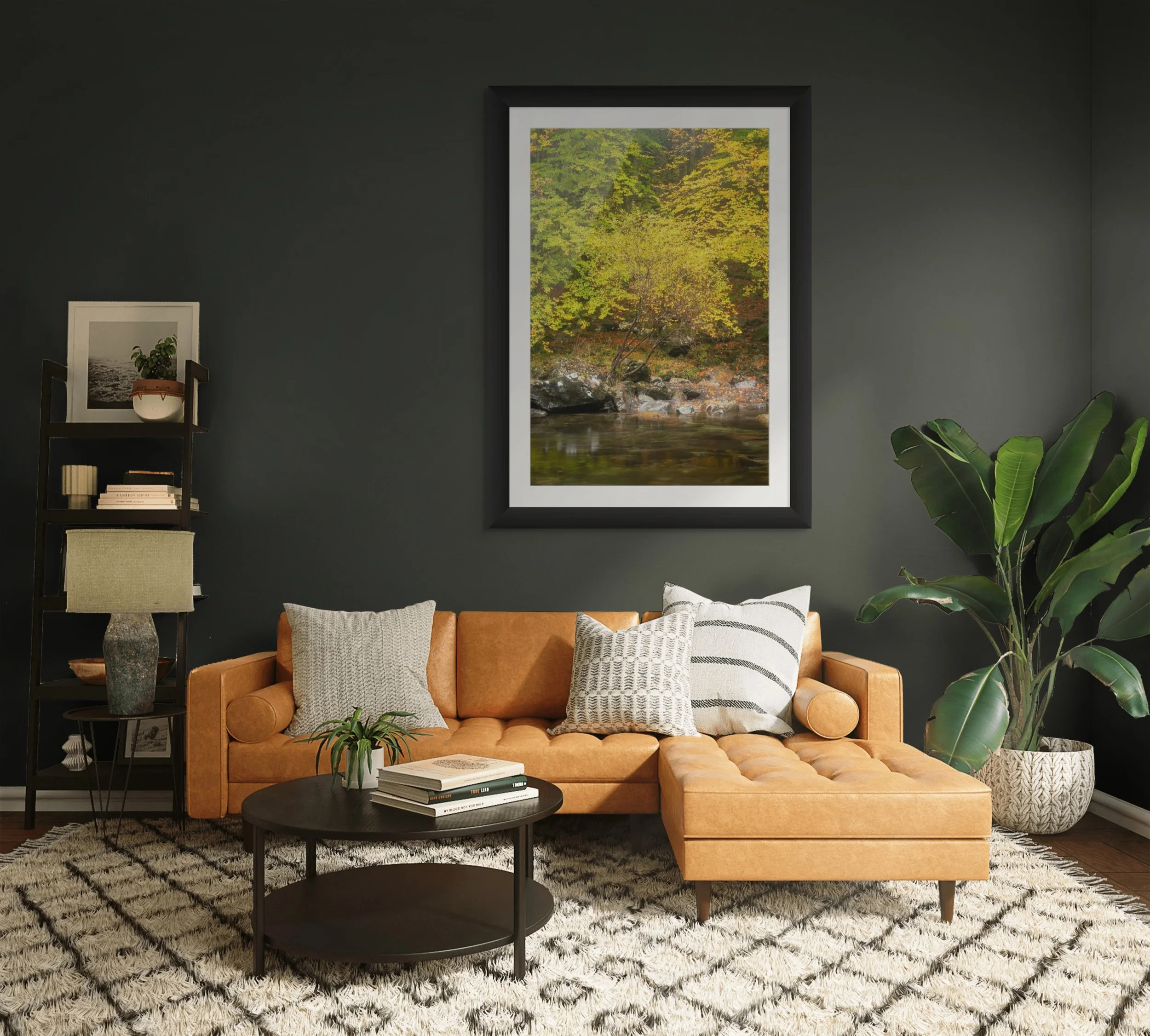 Living room with dark wall, framed forest landscape picture, black round coffee table with books, tan leather sofa with pillows, green plant, framed photograph, small shelves, and a lush potted plant.