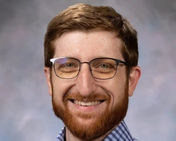 A smiling man with glasses, brown hair, and a beard, wearing a checkered shirt and a gray background.