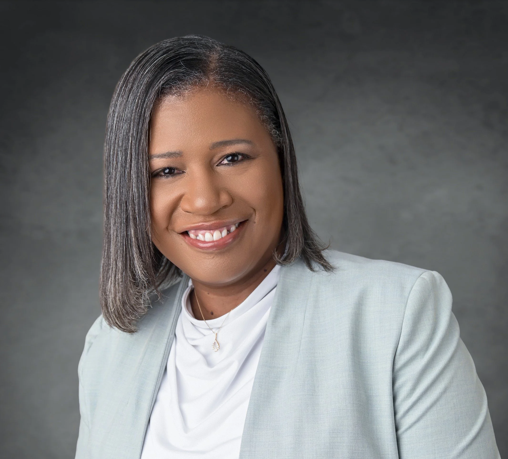 Close-up photo of a woman with shoulder-length, straight gray hair, smiling and wearing a light green blazer and white blouse, against a gray background.