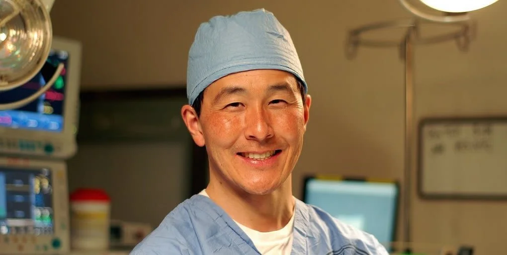 A smiling male surgeon in scrubs and surgical cap standing in an operating room.