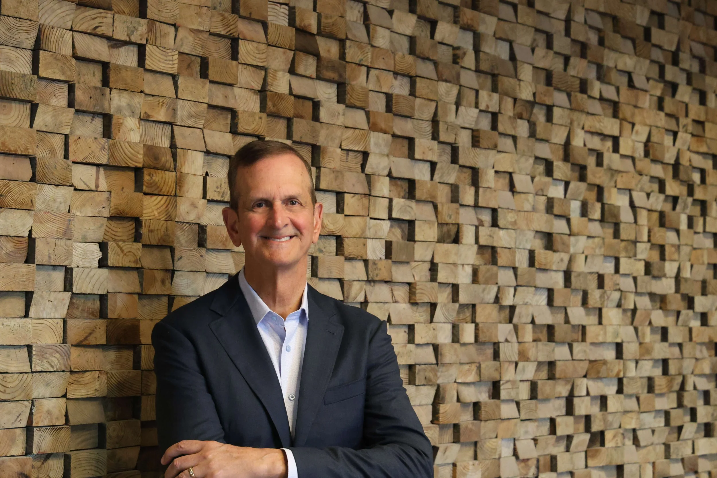 Law and accounting firm business development consultant Bob Silvy in a suit standing with arms crossed in front of a wall made of stacked wooden blocks.