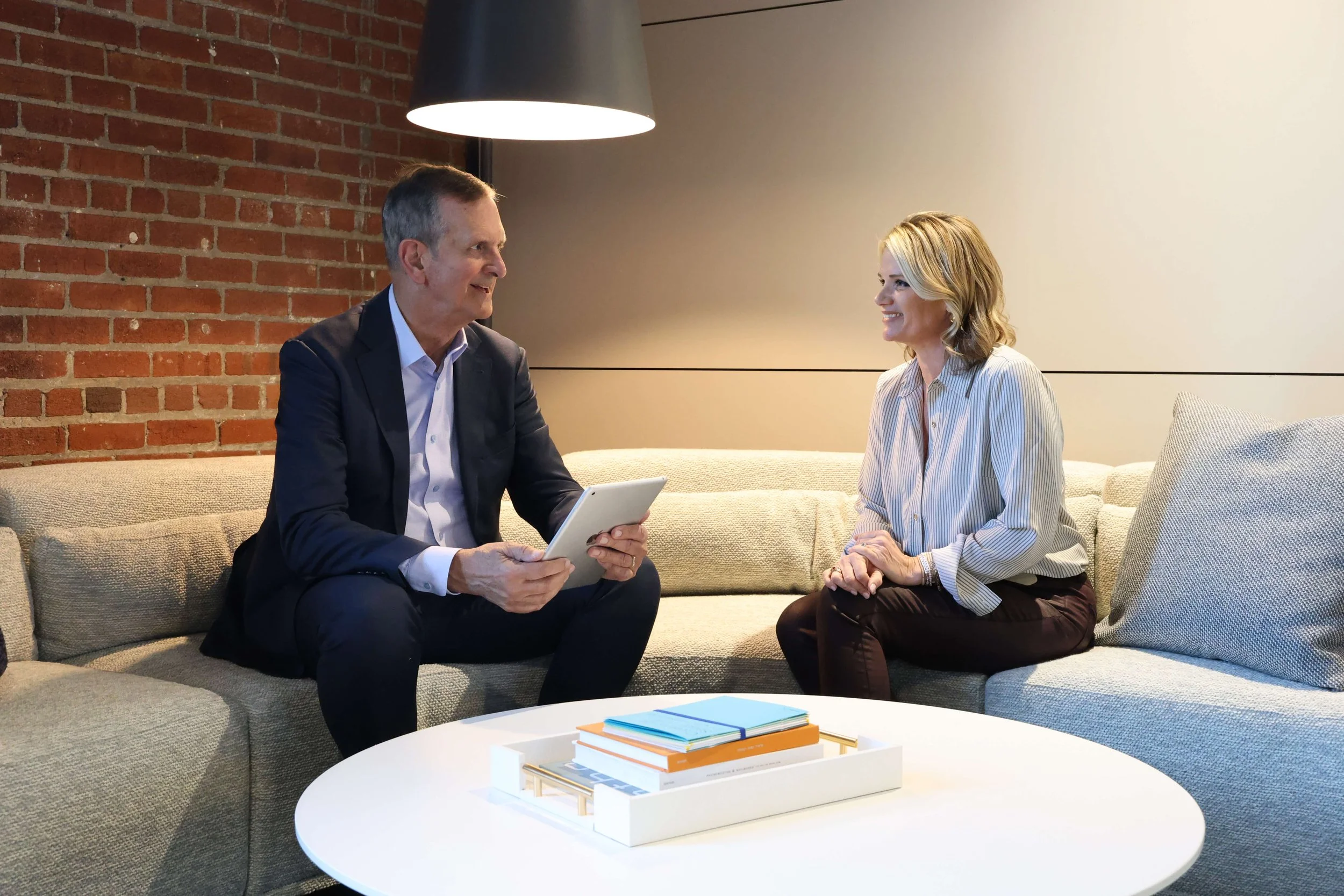 Law and accounting firm sales consultant Bob Silvy and client sit on a beige sofa in a modern, brick-walled room, engaged in conversation. Bob holding a tablet, and a white tray with colorful notebooks rests on a round white table in front of them.
