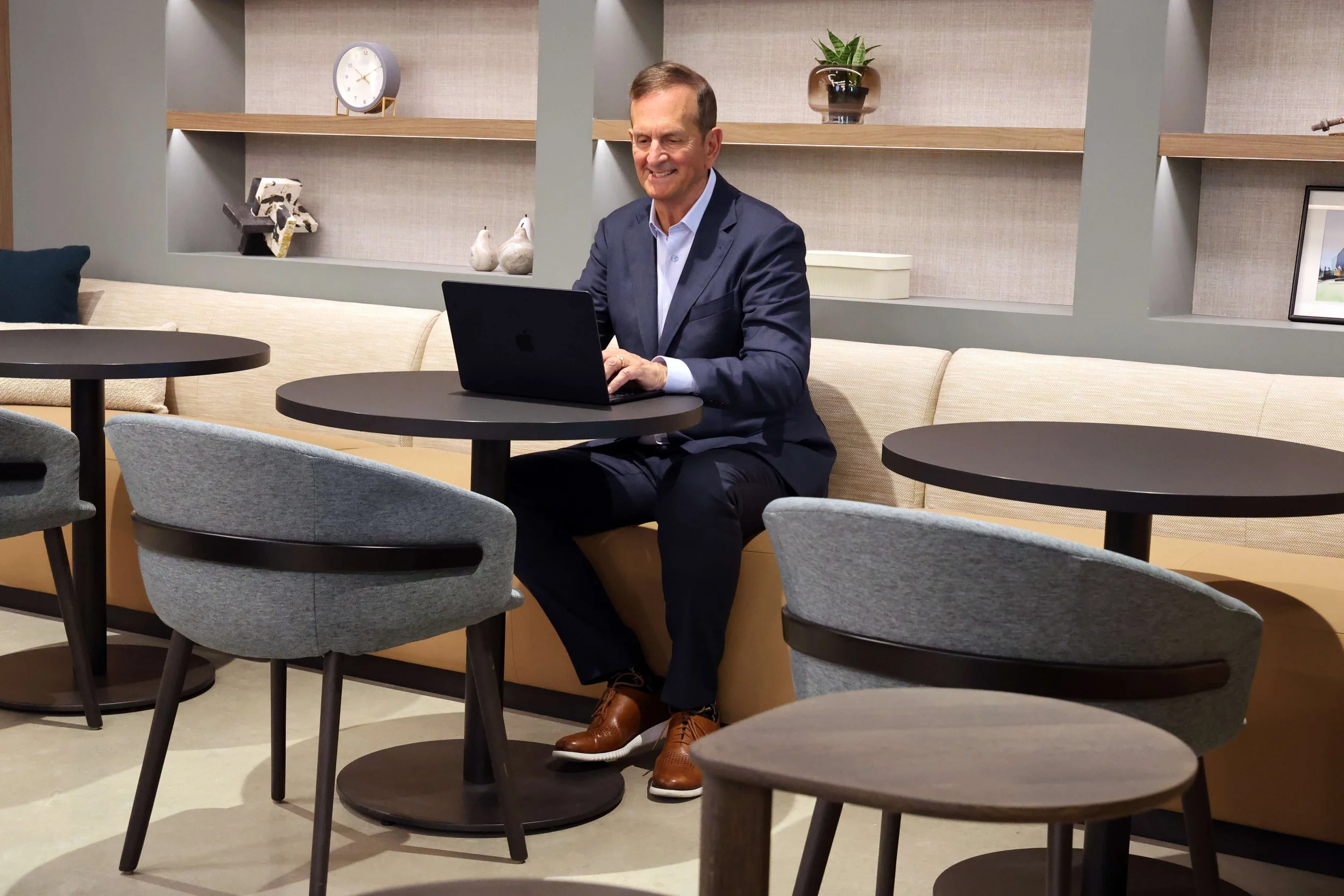 Law and accounting firm business development consultant Bob Silvy in formal attire working on a laptop in a modern lounge or lobby area with tables, chairs, and decorative objects on shelves.
