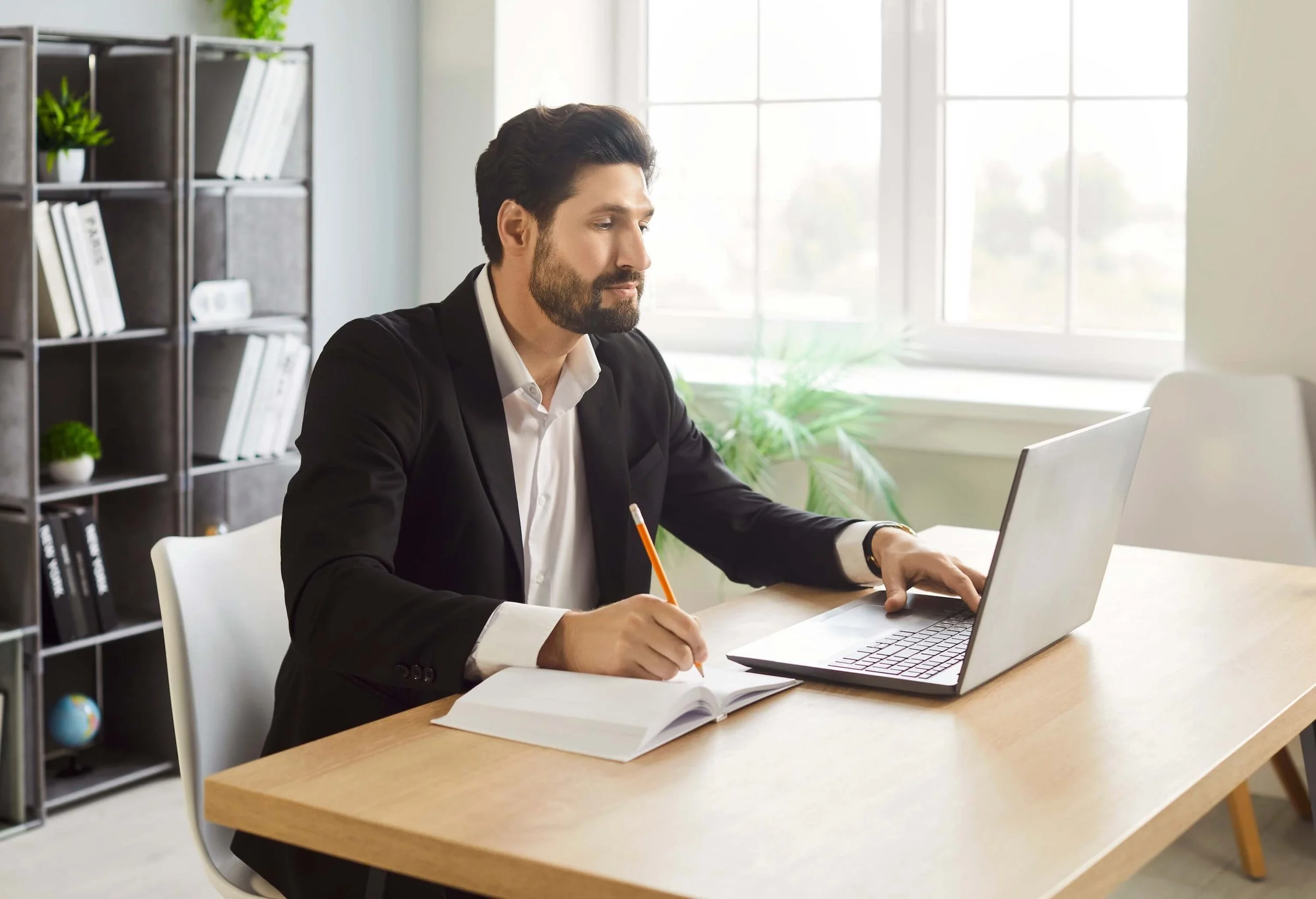 A man in a black suit and white shirt working at a wooden desk. He is holding a pencil in one hand and has an open book in front of him next to a silver laptop. Behind him is a bookshelf with plants, books, and decorative items, and a window letting in natural light.