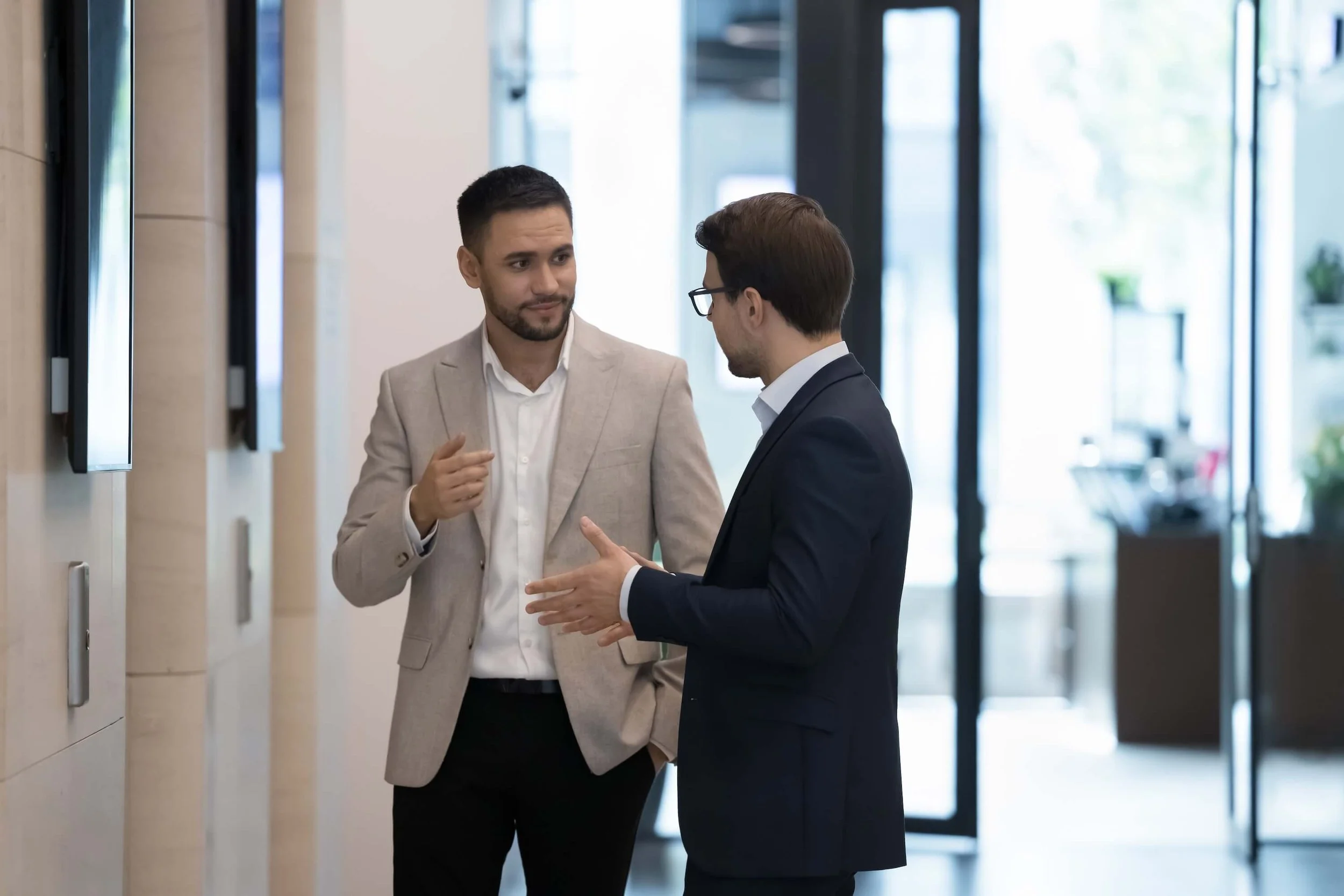 Two men engaged in conversation in a modern office lobby, one wearing a light beige suit and white shirt, and the other in a dark navy suit and glasses, with a glass door and windows in the background.
