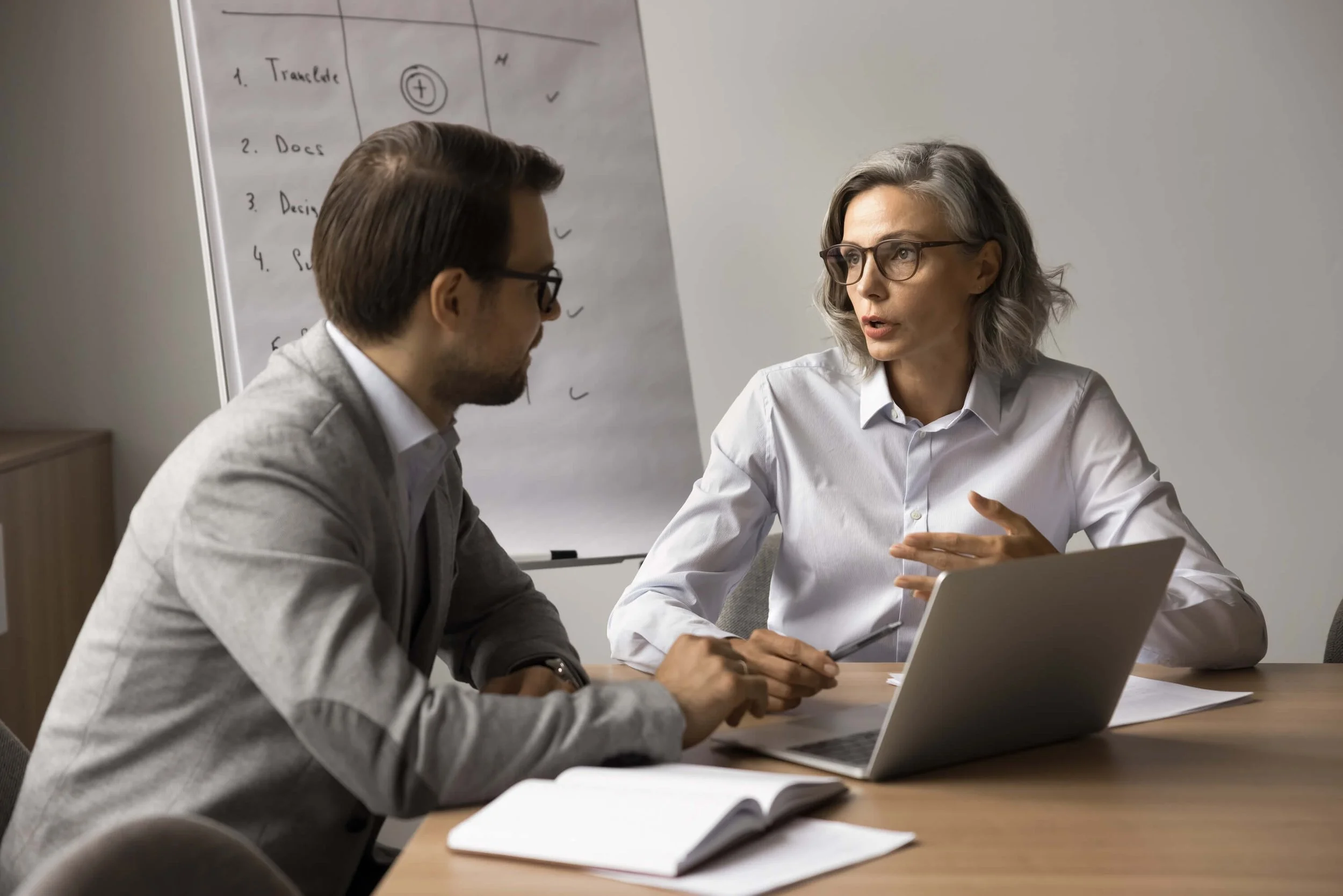 A woman and a man having a serious discussion in a meeting room, with a whiteboard filled with notes and checkmarks in the background.