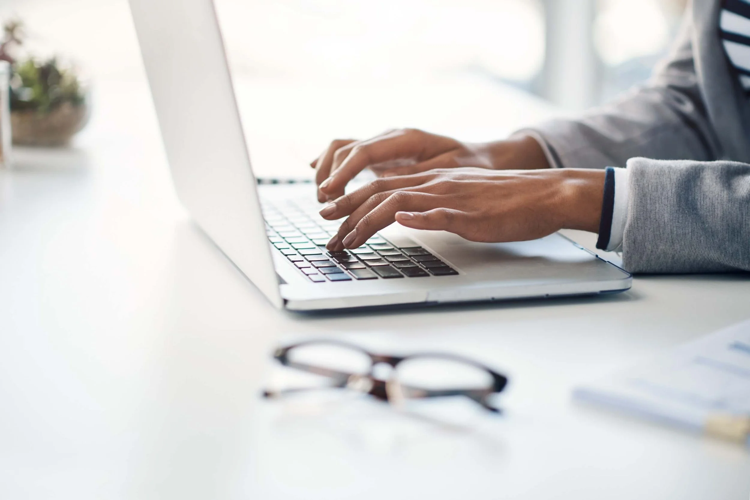 Person typing on a laptop at a white desk with glasses and papers.