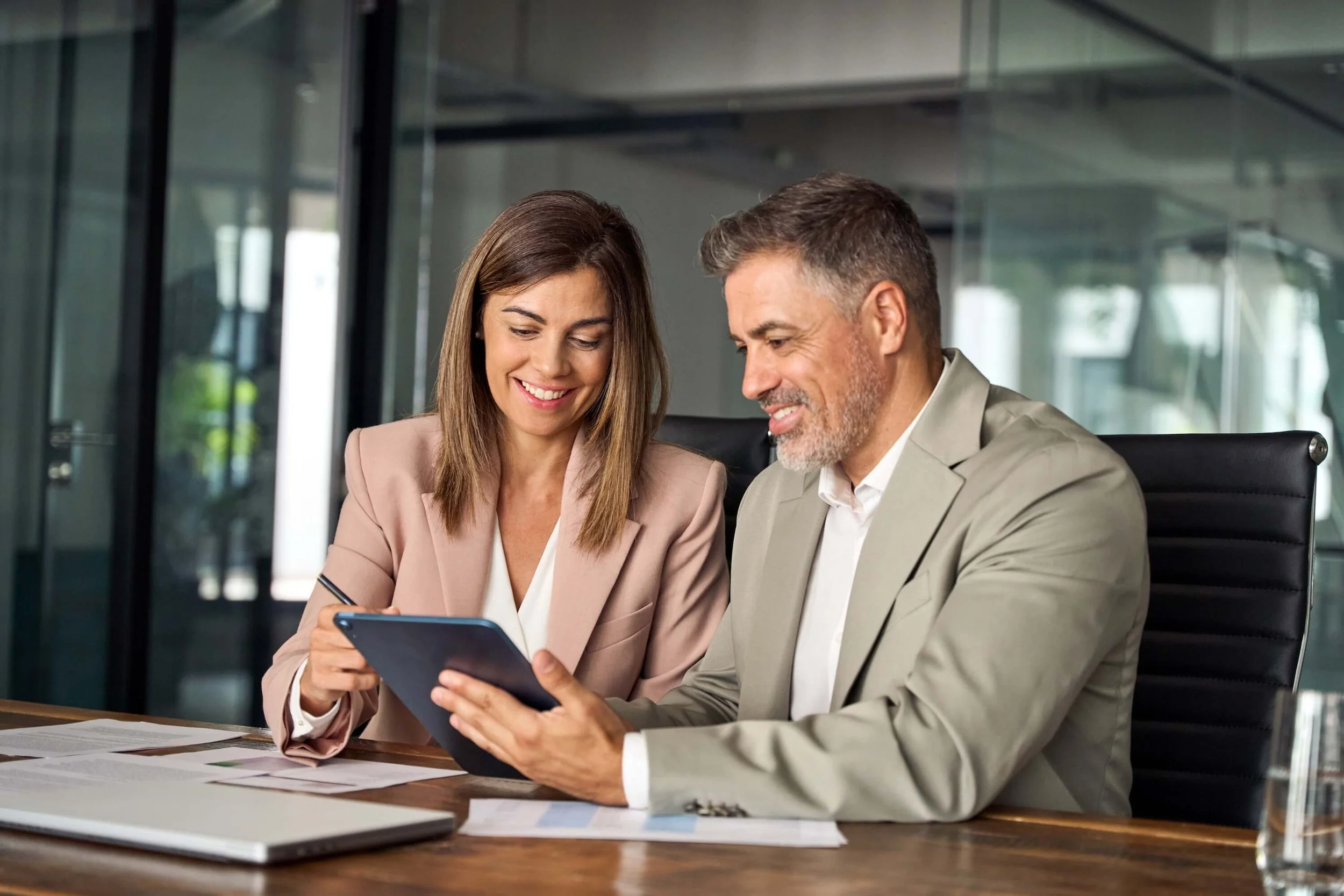 A man and woman in business attire sitting at a desk in an office, looking at a tablet together and smiling.