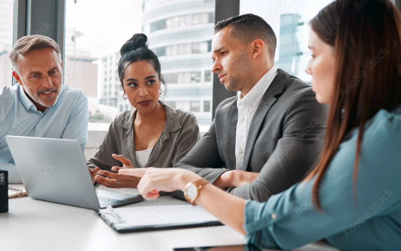 A diverse group of five professionals in a business meeting around a table in an office with large windows and urban buildings in the background.