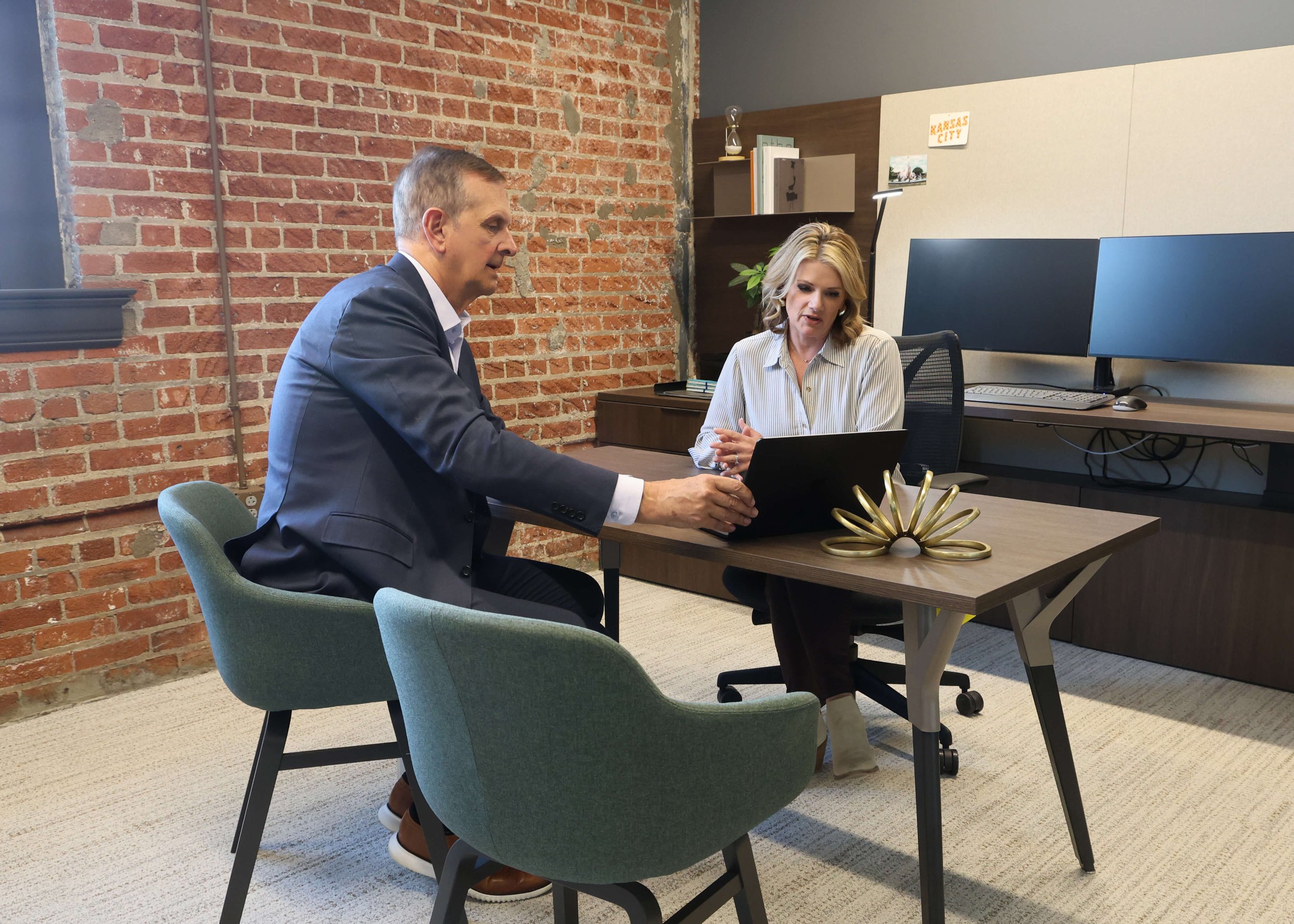 Two professionals, a man and a woman, sitting at a desk in an office engaged in a discussion over a laptop, with the man pointing at the laptop screen.