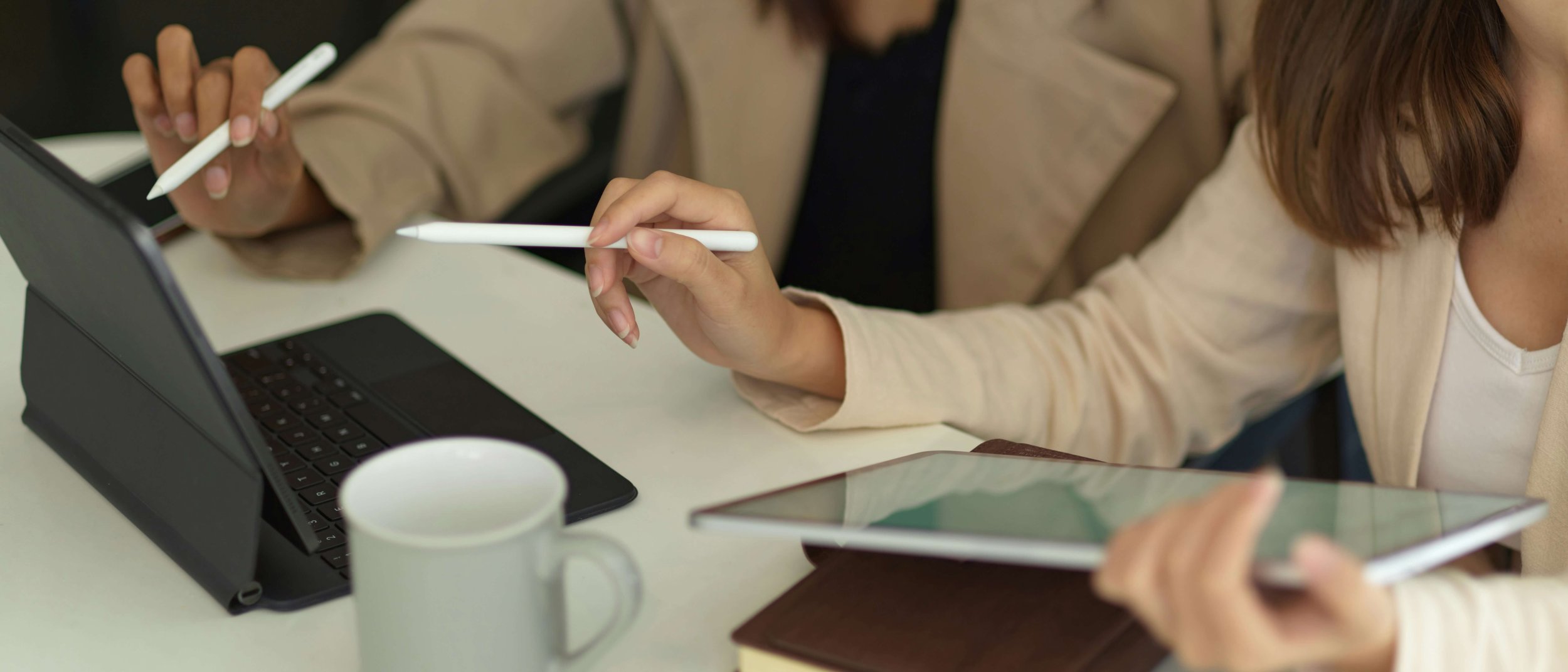 Two women working at a desk with digital tablets, a laptop, and a coffee mug.