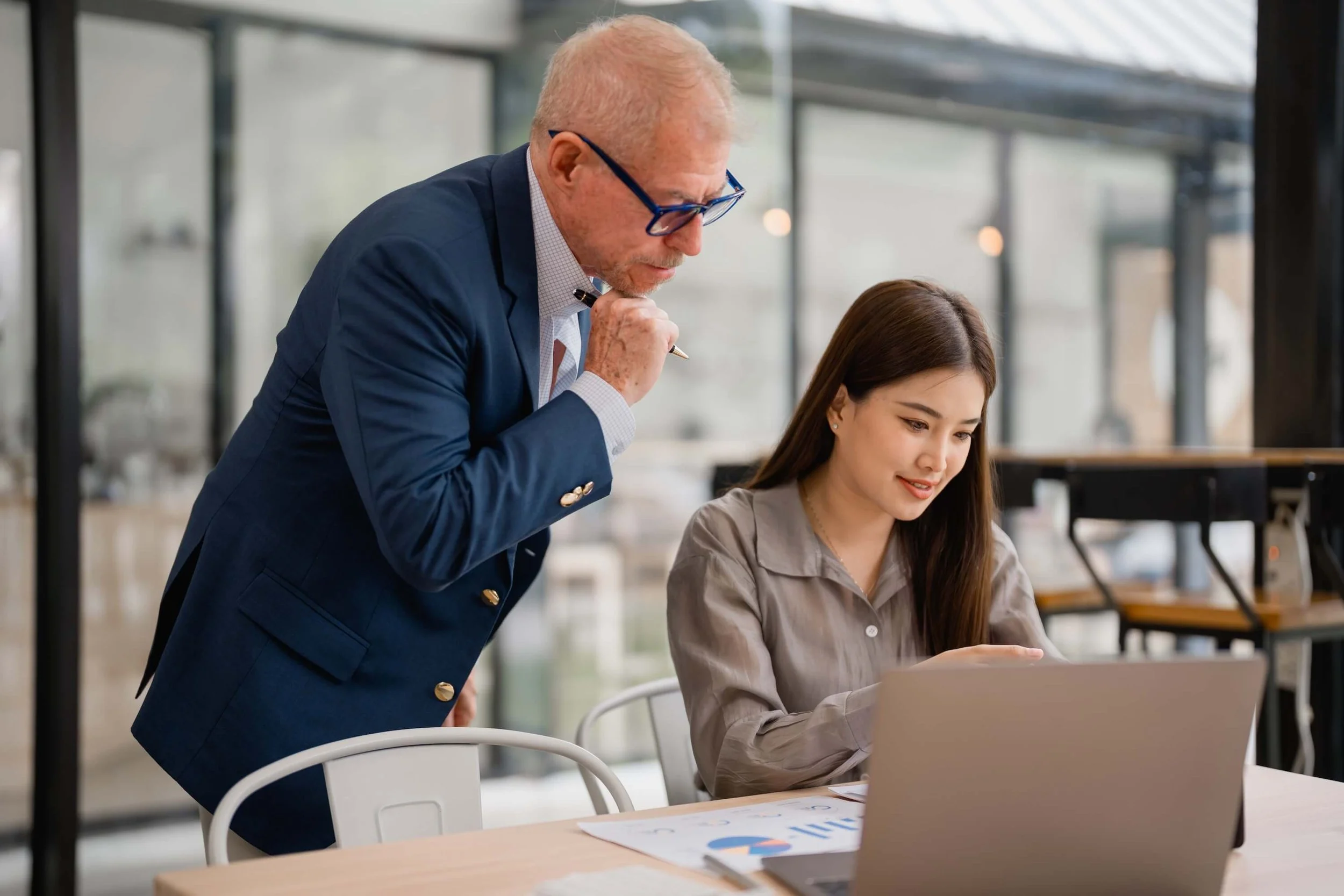 A man in a blue suit and glasses leaning over to look at a laptop screen held by a woman with long brown hair, sitting at a wooden table in a modern office space with large windows.