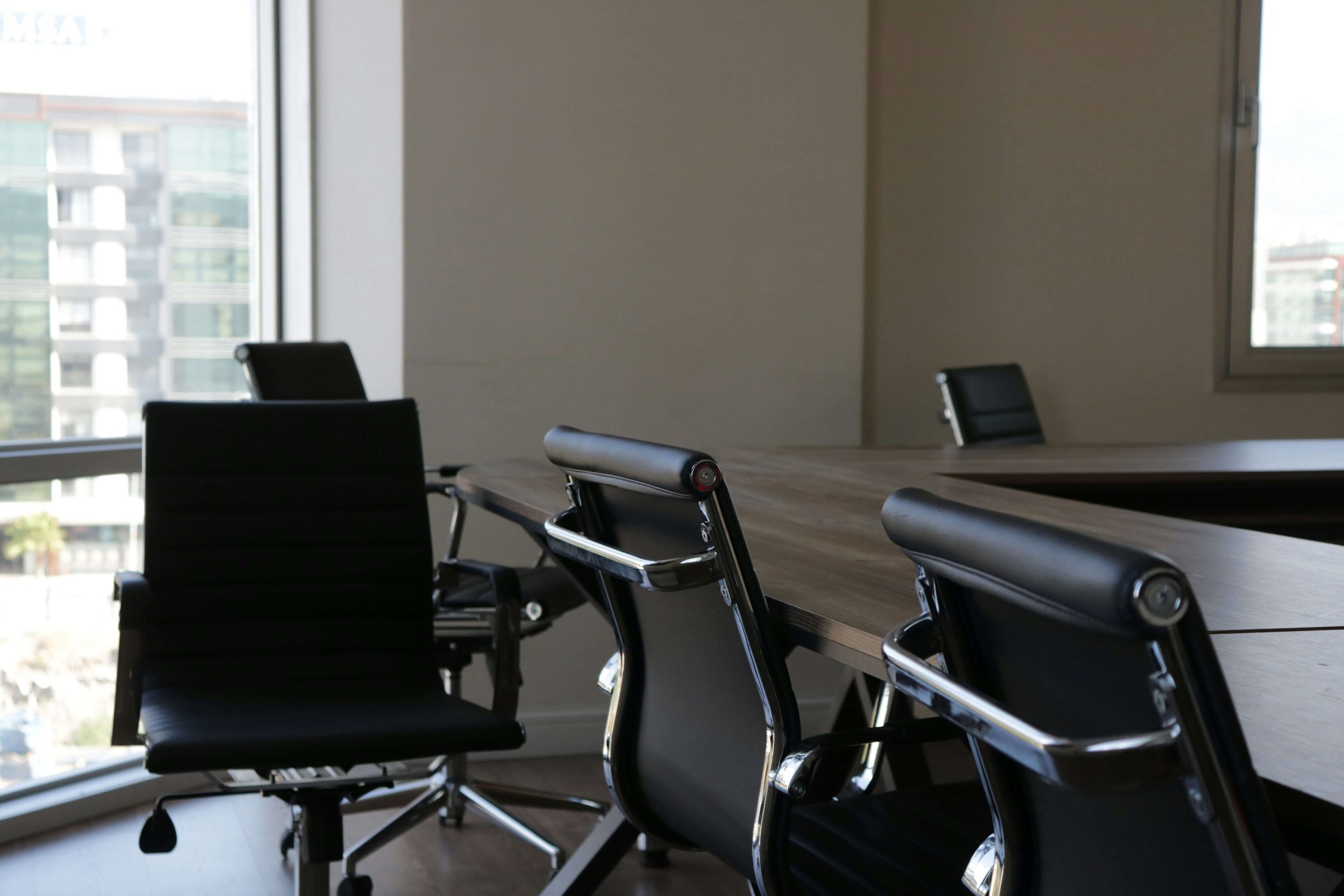 Empty conference room with a large wooden table and black office chairs, large windows showing city buildings, natural light