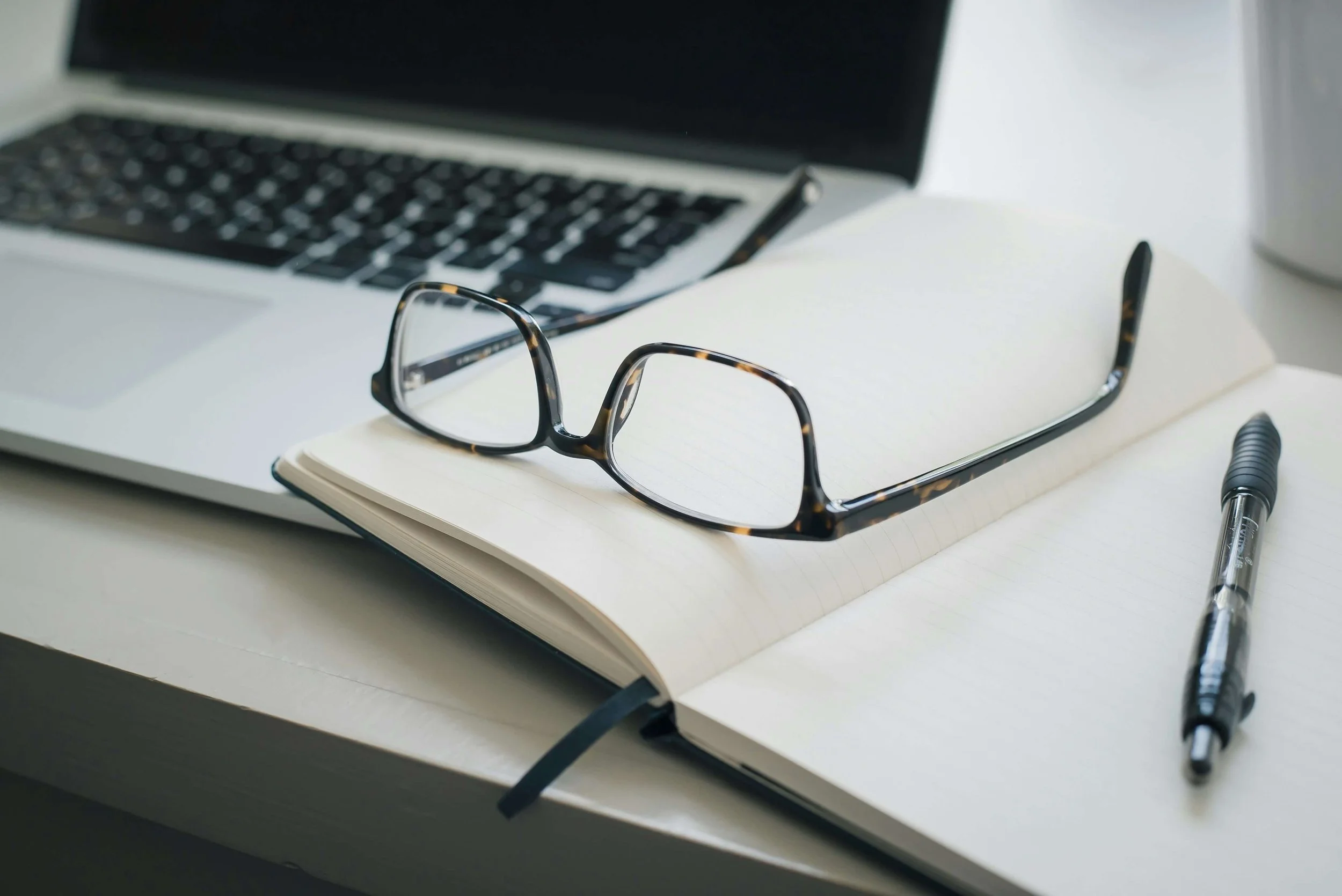 A pair of tortoiseshell eyeglasses resting on an open notebook with blank pages, a black pen beside it, and a laptop in the background on a white desk.