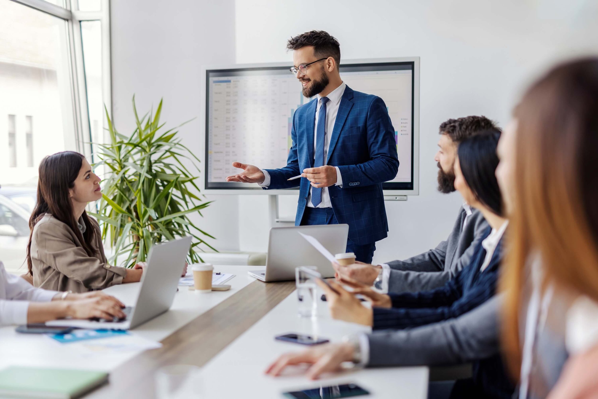Business meeting with a man in a blue suit speaking to a group in a conference room with laptops and a large screen display.