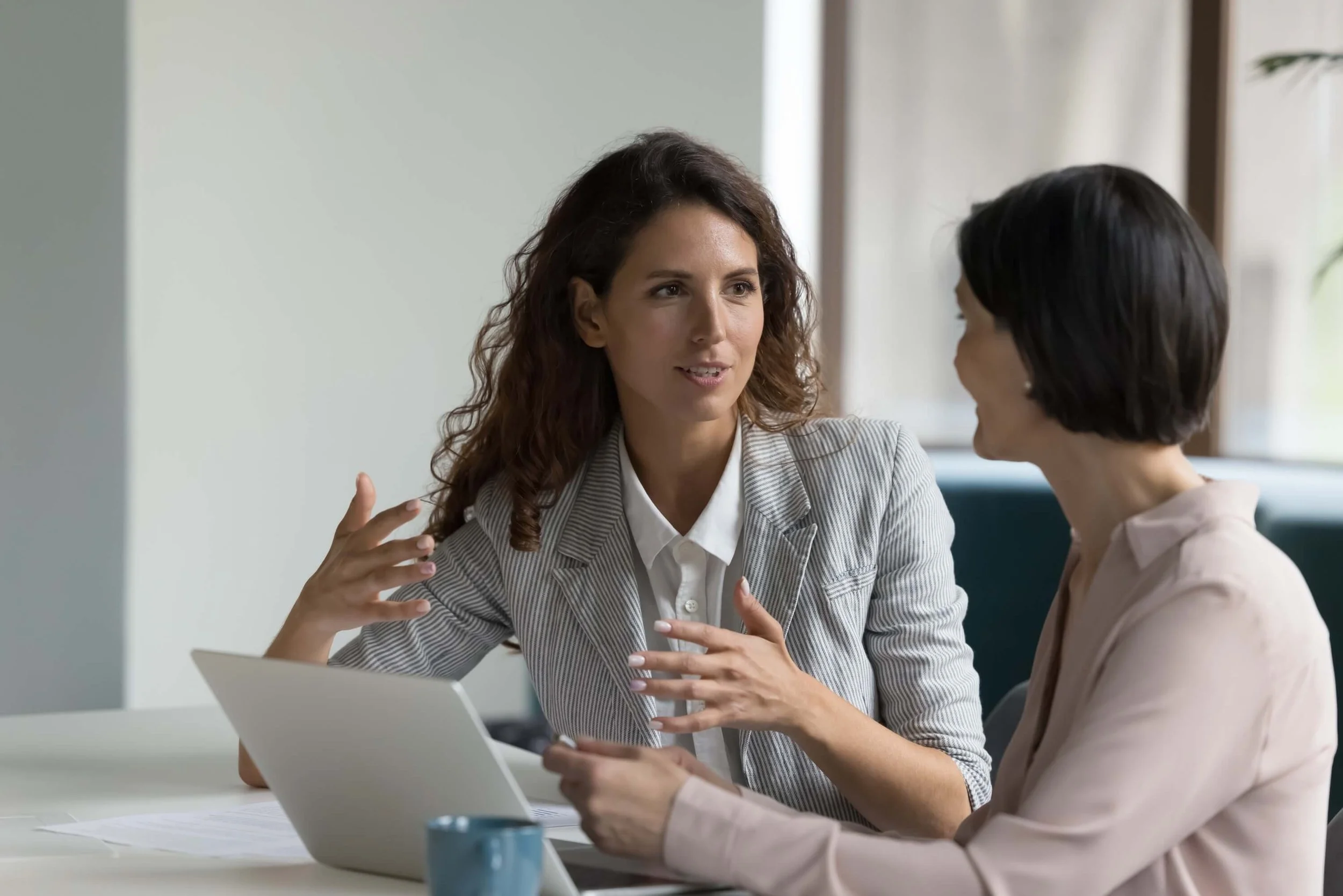 Two women having a conversation at a table with a laptop and a mug in front of them, in a bright, modern office space.