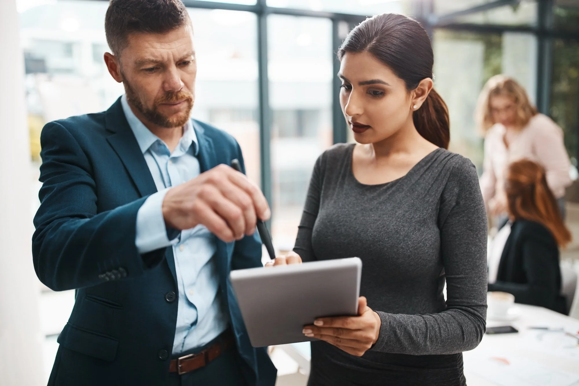A man and woman in business attire looking at a tablet together in an office setting, with two women working in the background.