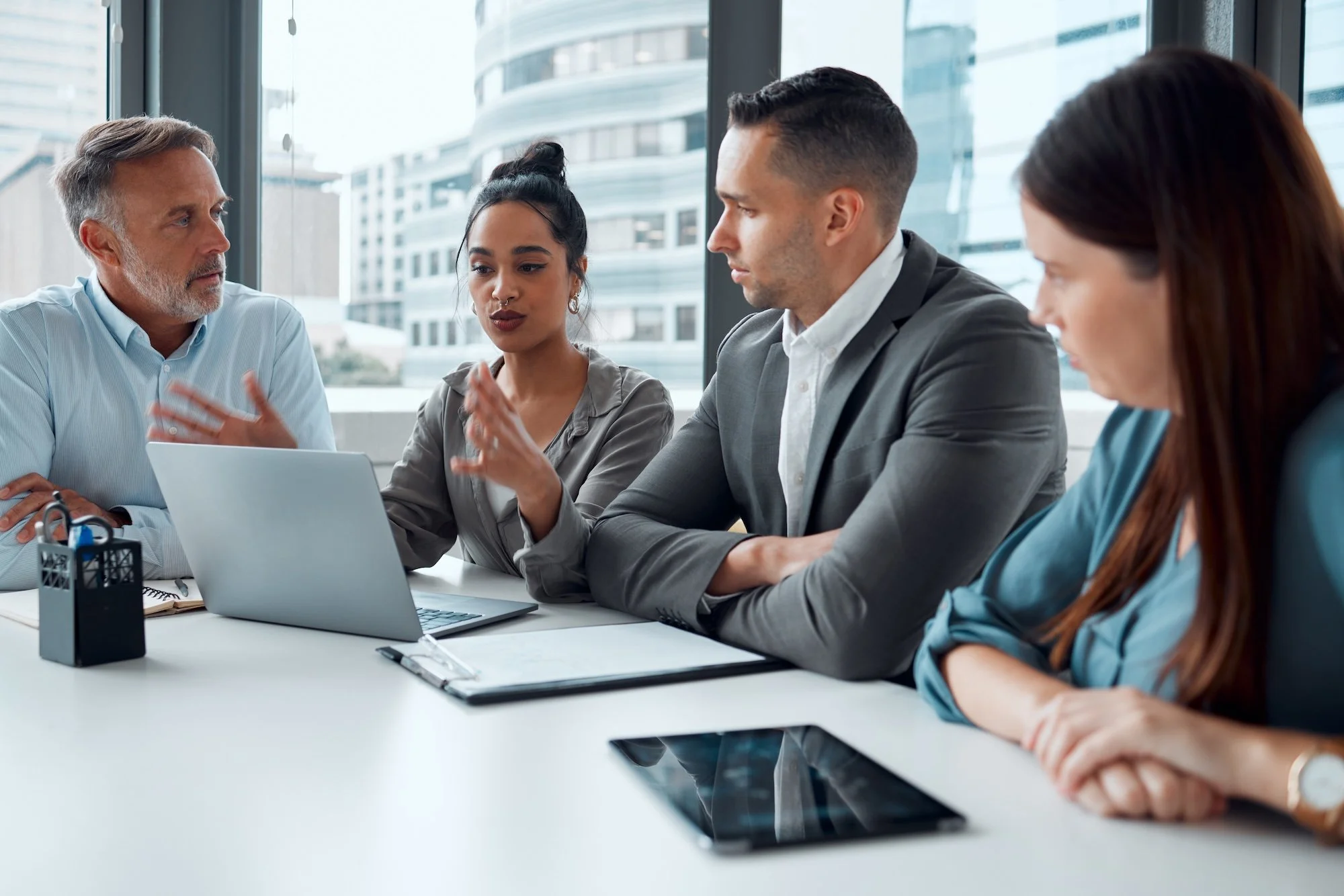 Five professionals in a meeting room with large windows showing city buildings, engaged in discussion around a table with laptops, tablets, notebooks, and office supplies.