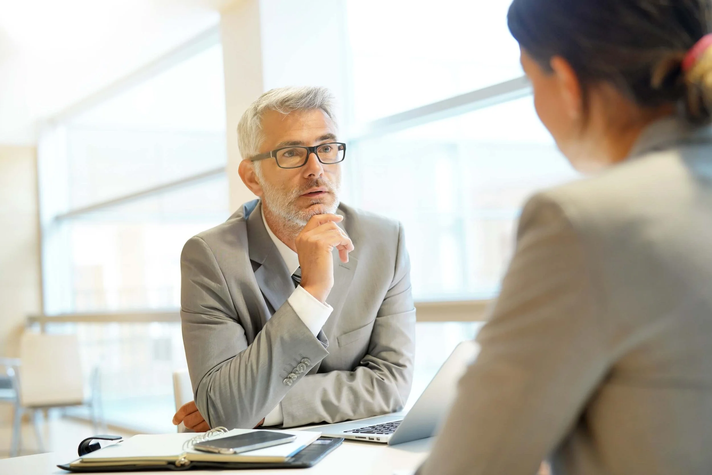 Two professionals in a business meeting; the man with gray hair and glasses is speaking, the woman with dark hair tied back listens attentively.