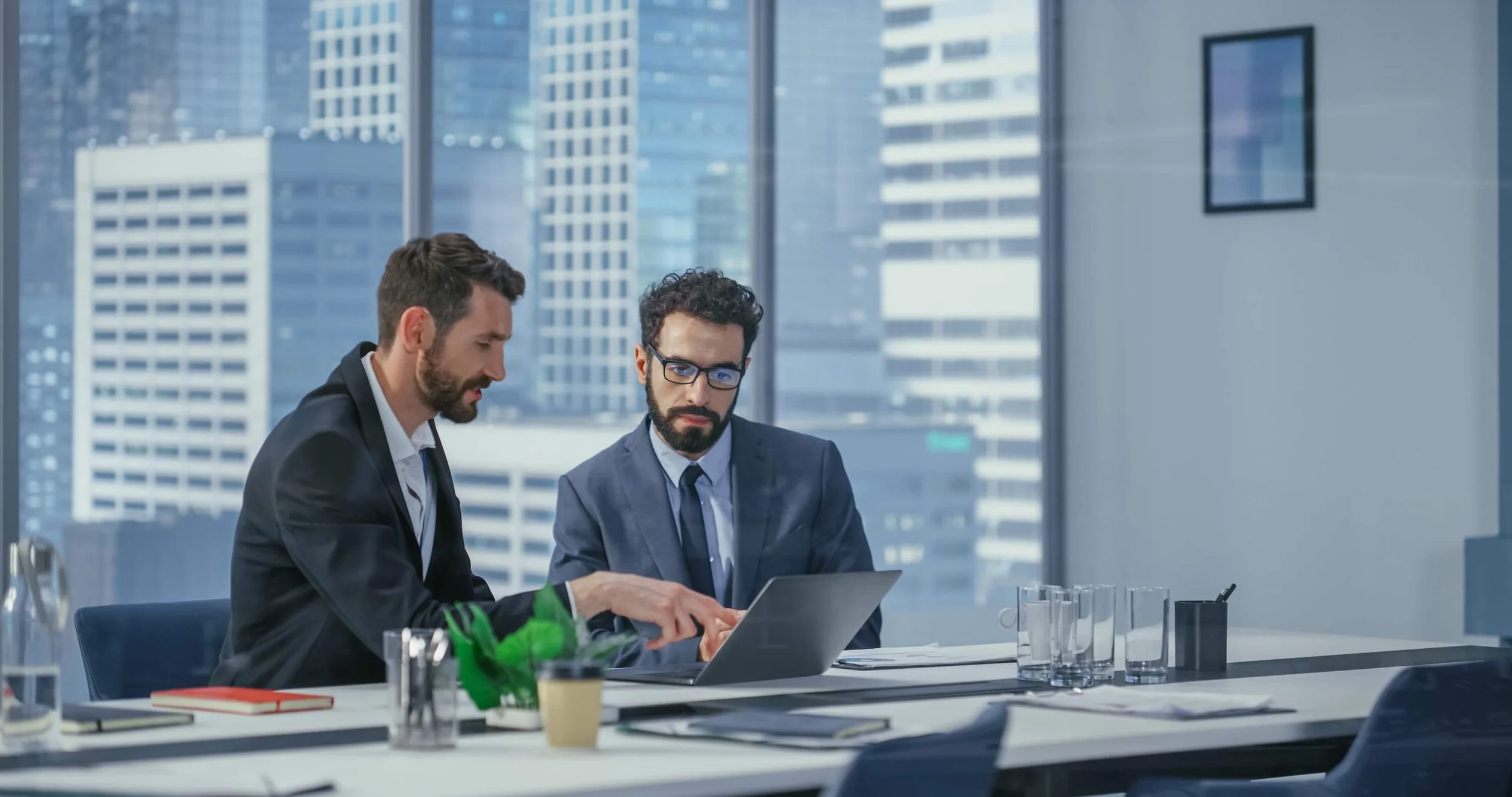 Two businessmen in suits working together on a laptop at a conference table in a modern office with large windows and cityscape view.
