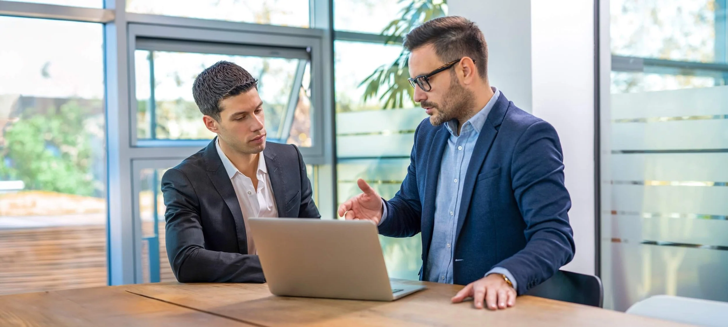 Two men in business suits having a discussion over a laptop in a modern office with large windows and natural light.