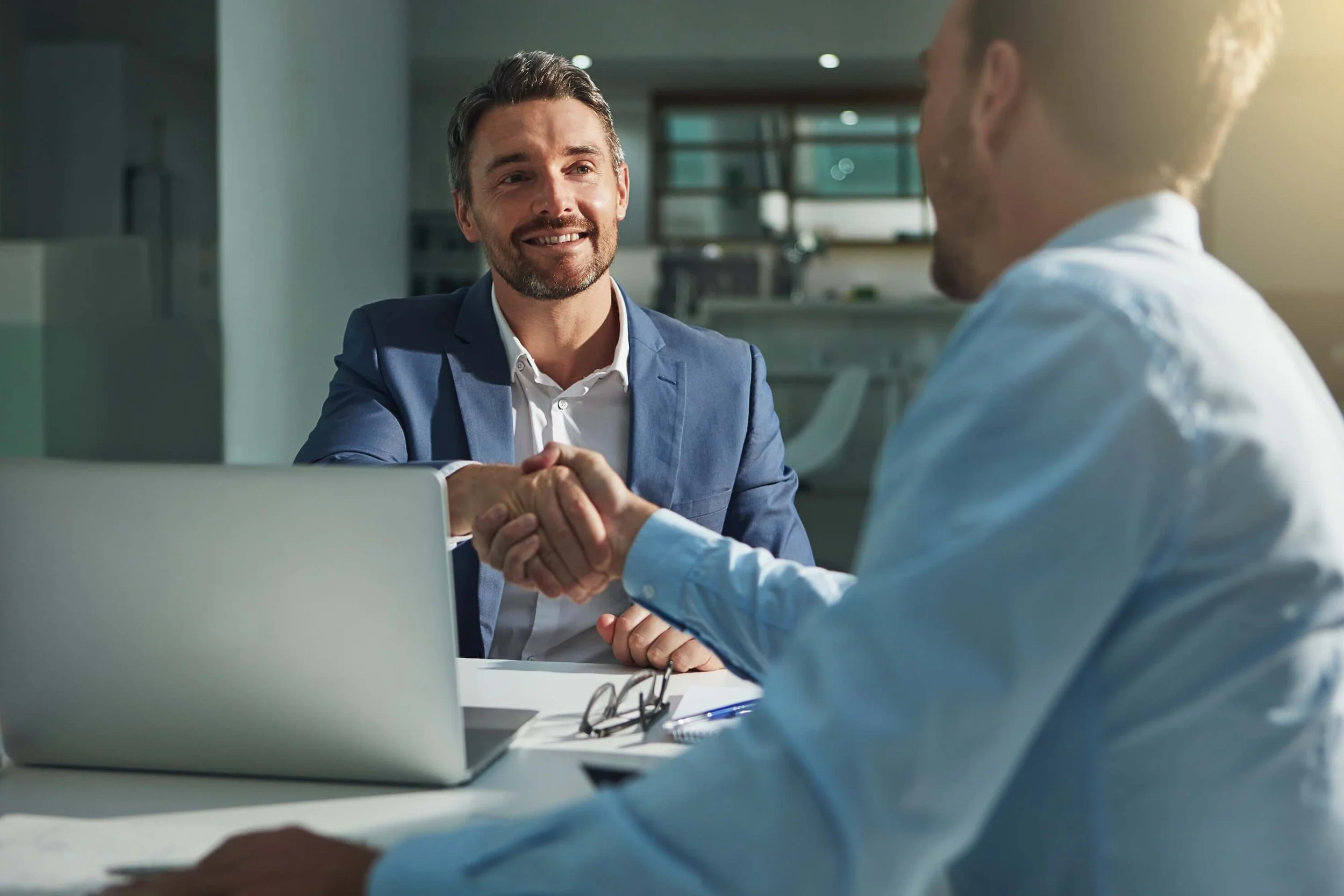 Two men in business attire shaking hands during a meeting in an office, with a laptop, notepad, and glasses on the table.