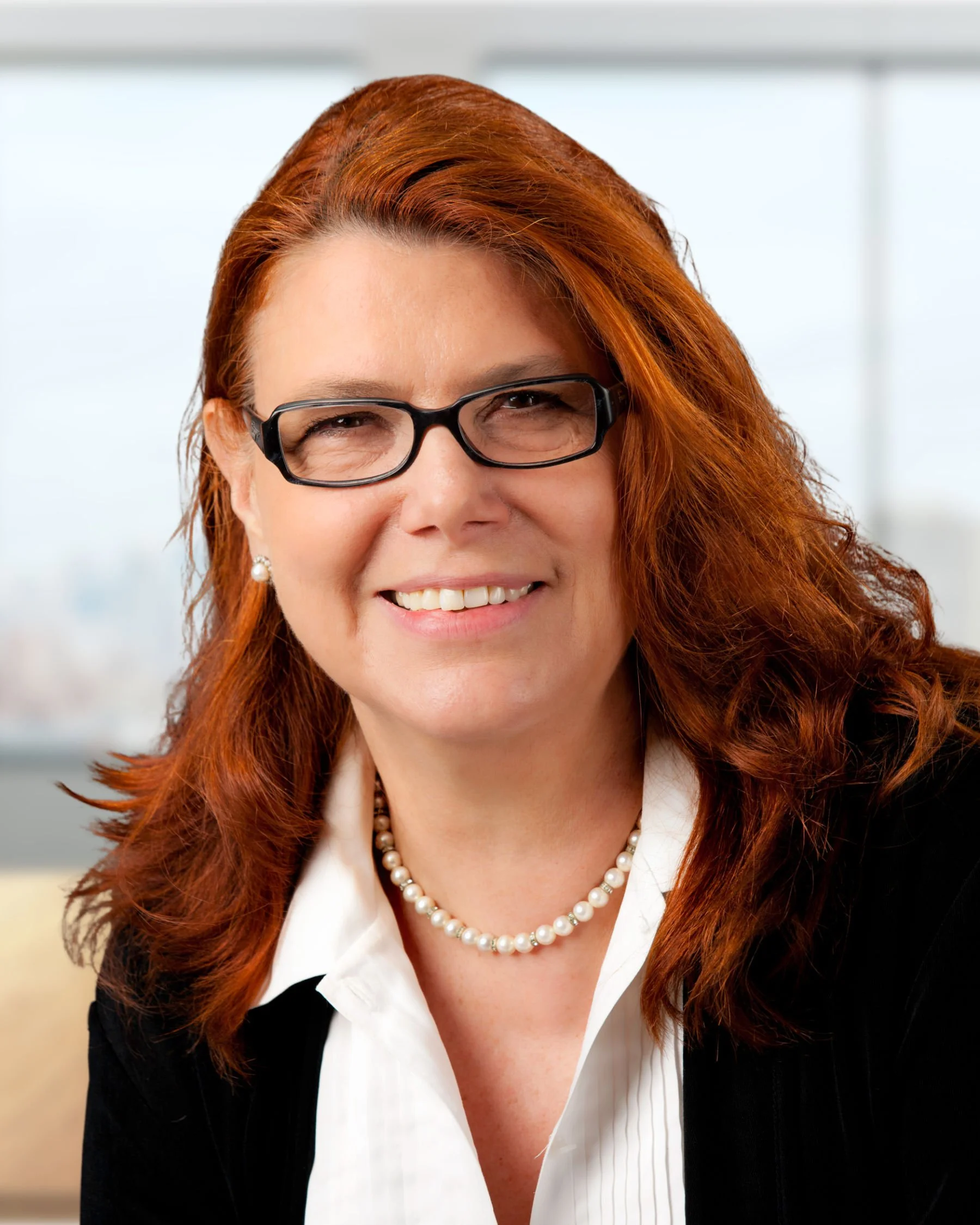 Portrait of a smiling red-haired woman wearing glasses, a pearl necklace, a white shirt, and a black blazer, in an office setting.