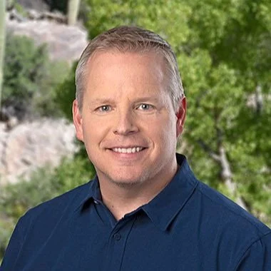 A smiling man with short light-colored hair, wearing a dark blue button-up shirt, standing outdoors with green trees and rocks in the background.