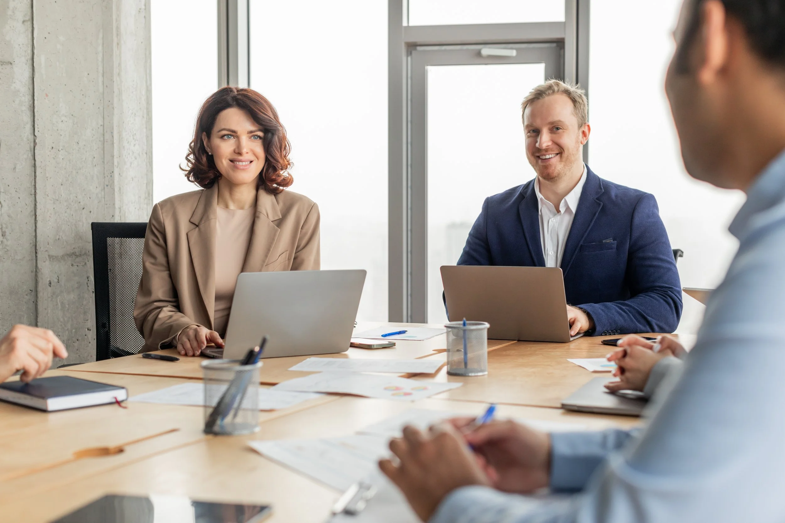 Business meeting with four people in a conference room, two of whom are clearly visible, smiling and looking towards the person speaking, with laptops and documents on the table.
