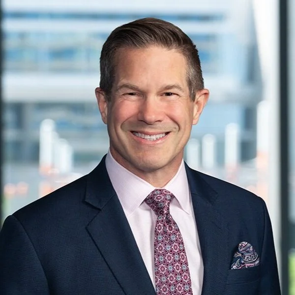 A smiling man in a navy suit, light pink shirt, patterned tie, and pocket square, standing in front of a blurred cityscape window background.