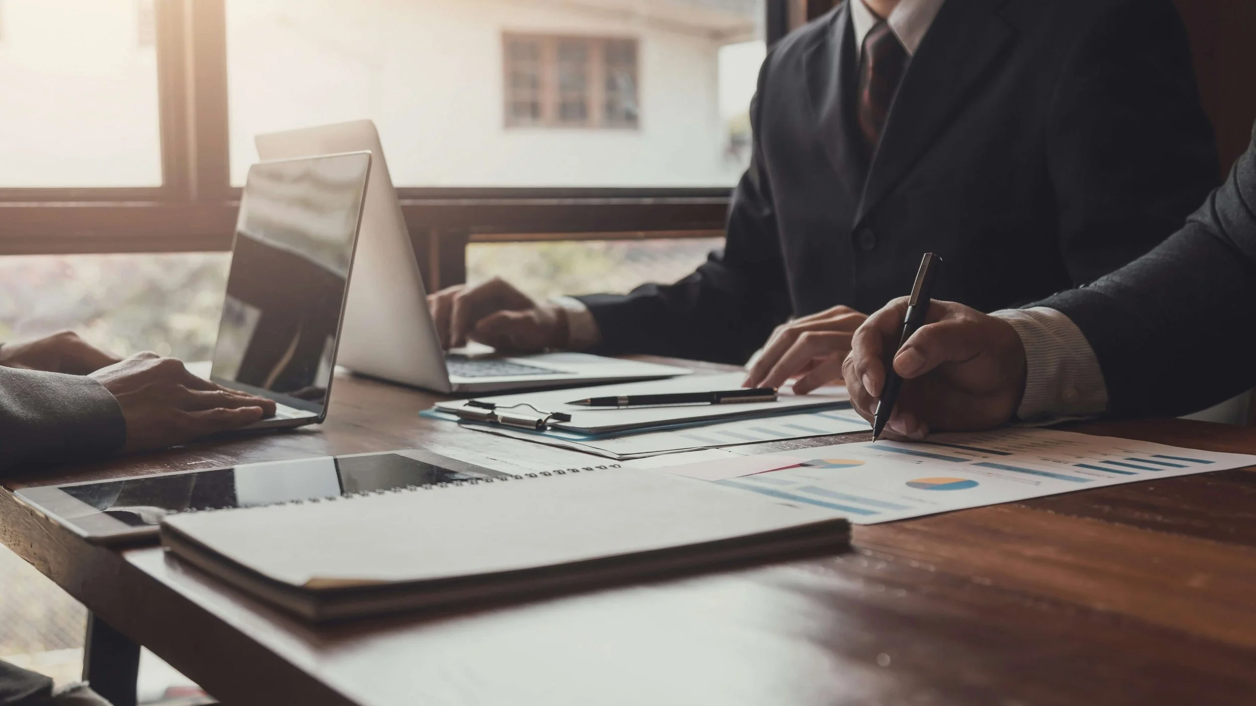 Business professionals in suits reviewing financial documents and graphs on a table with laptops and notebooks in a conference room with natural light.