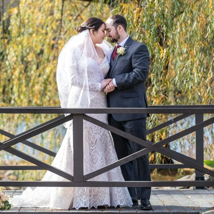 Bride and groom on a wooden bridge, touching foreheads, with trees in the background.