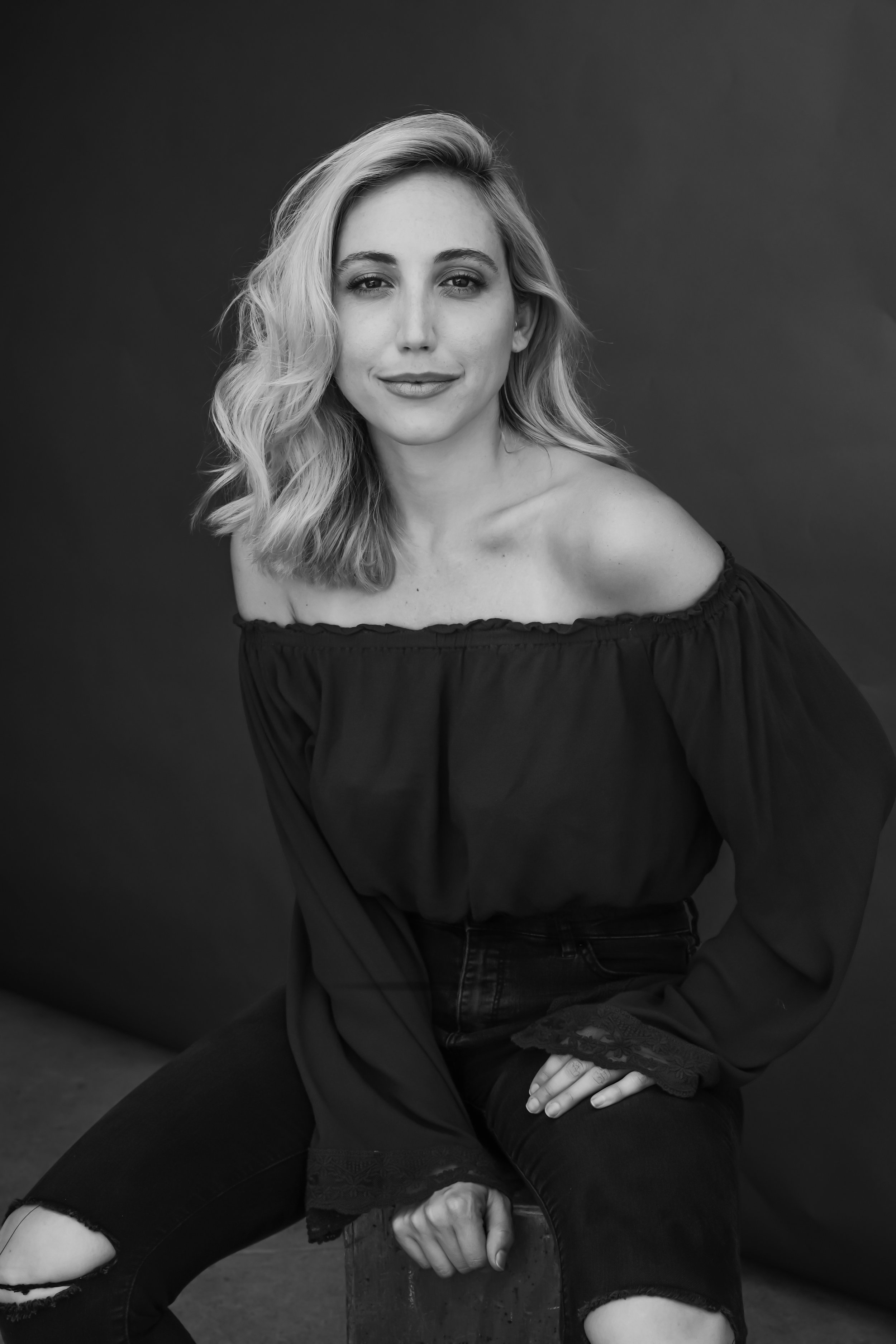 Black and white portrait of a woman with wavy blonde hair, smiling slightly, wearing an off-the-shoulder black top and ripped jeans, sitting on a stool against a dark background.
