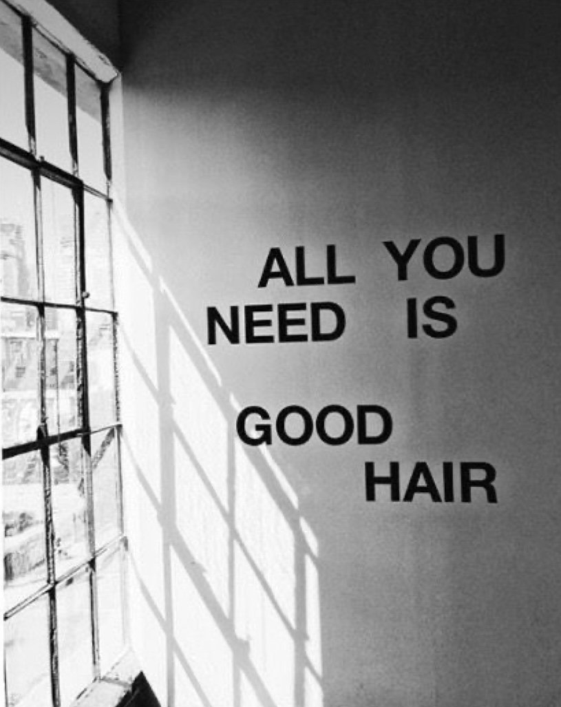 A black and white photo of a wall with large text that reads, "ALL YOU NEED IS GOOD HAIR," next to a window with grid panes casting shadows on the wall.