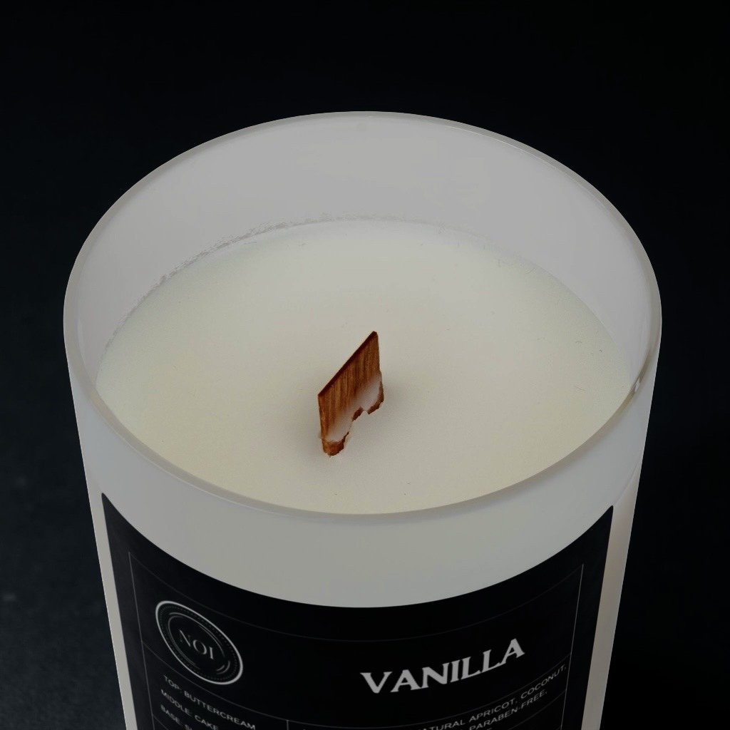 A white vanilla scented candle with a wooden wick in a black container labeled "Vanilla".