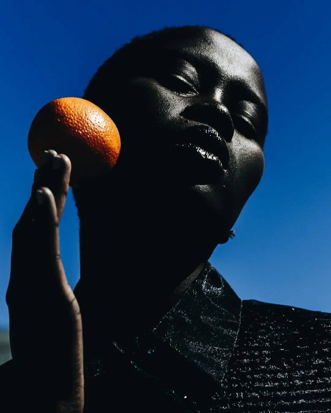 A close-up of a person with dark skin holding an orange next to their face against a clear blue sky.
