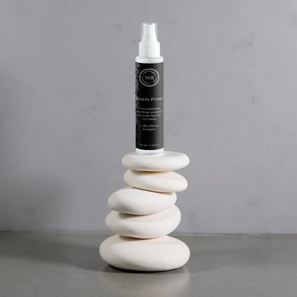 Keratin primer bottle stands on a tower of 5 white rounded rocks with an off white background and a neutral glossy stone floor