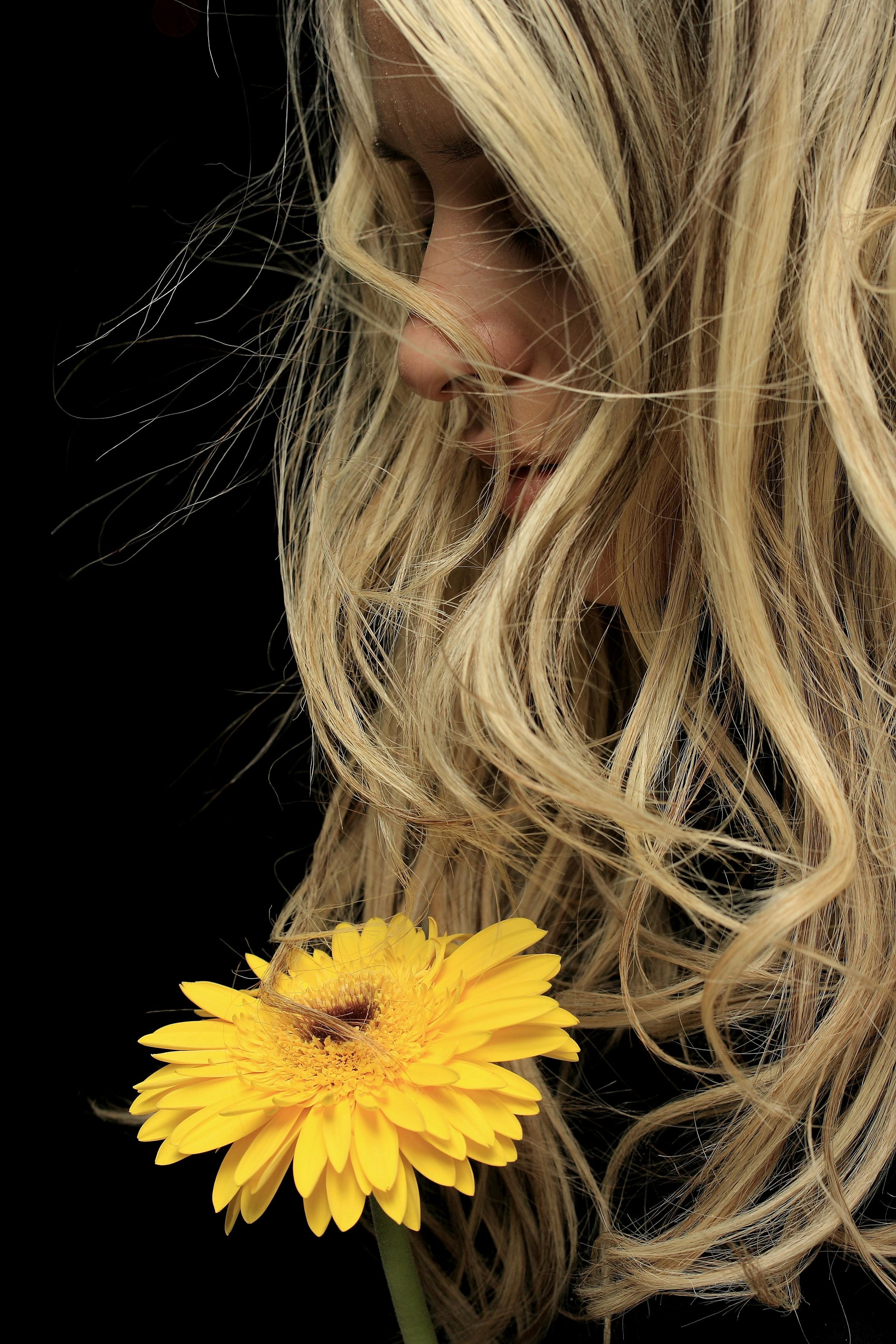A woman with long, wavy blonde hair looking down at a yellow flower against a black background.